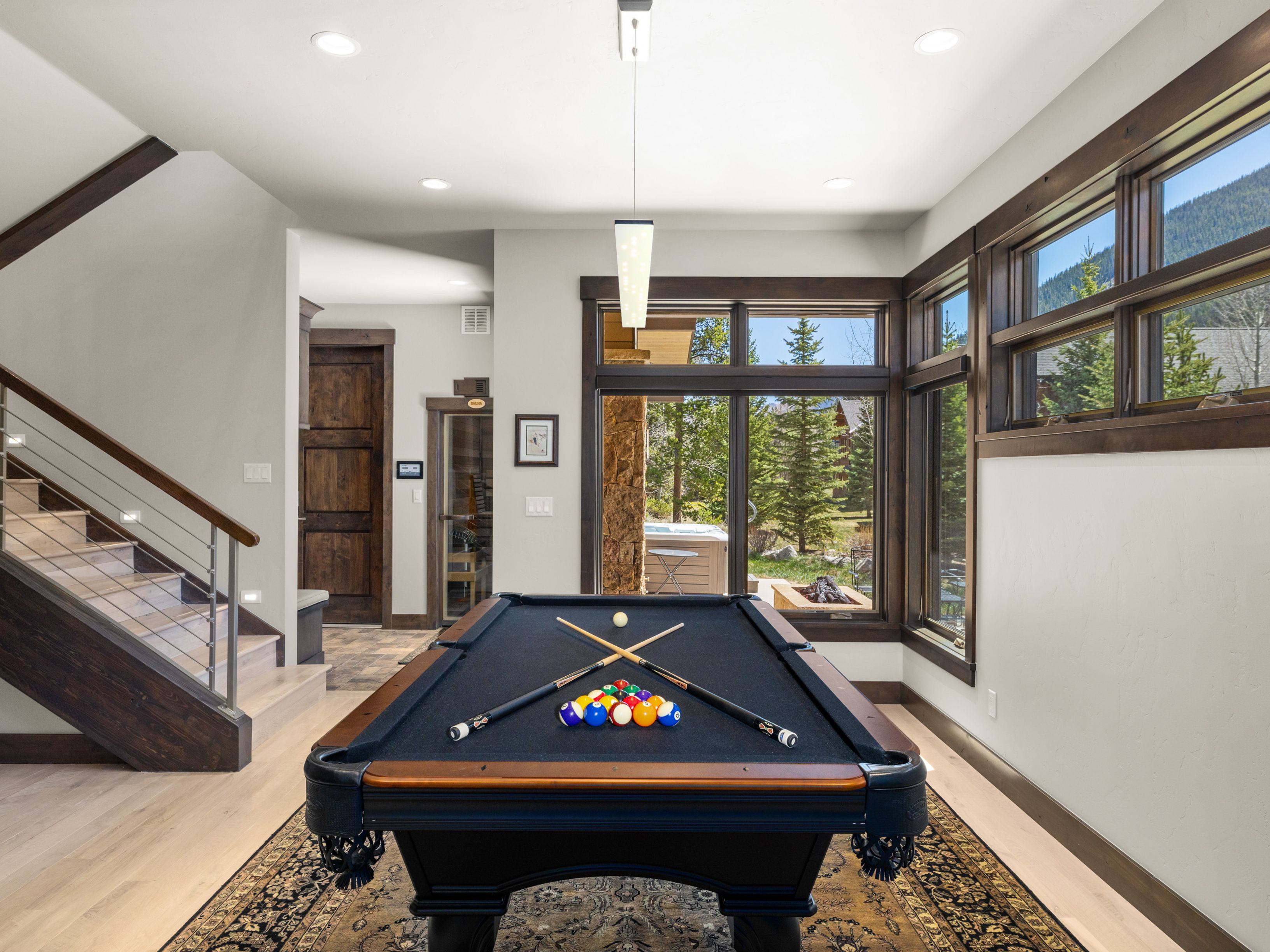 This interior space features a prominent pool table as the central focus, set upon a detailed patterned rug. To the left, a staircase with light wood treads and a modern metal railing leads upward, while large windows to the right provide a view of the outdoor landscape. The room is finished with neutral walls and dark wood trim, creating a sophisticated and inviting game room atmosphere.