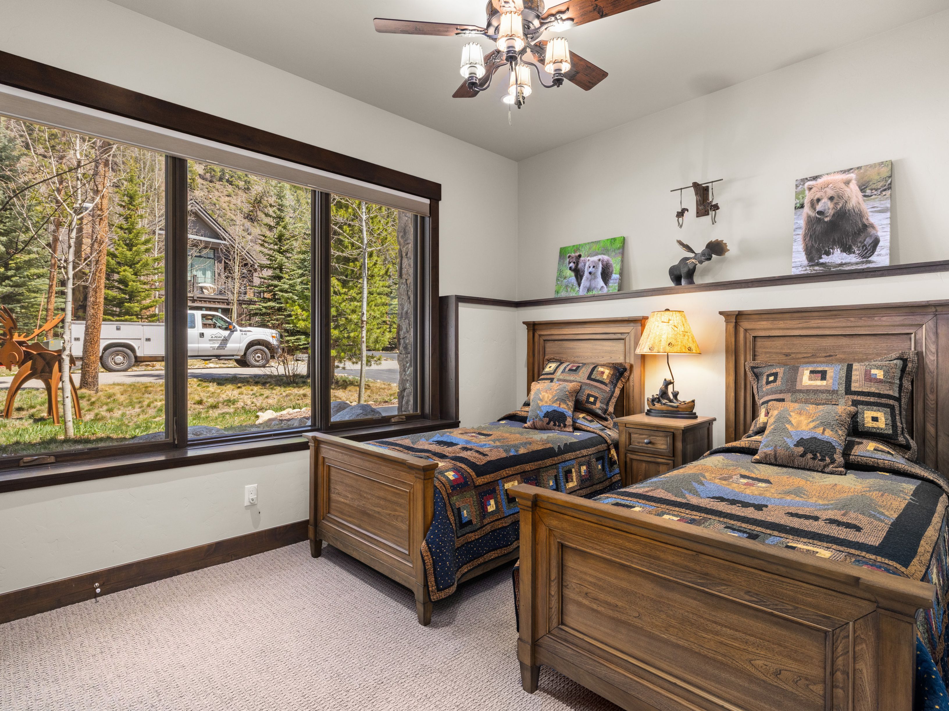 This cozy guest bedroom features two twin beds with rustic, bear-themed patterned bedding and wooden frames. A large window provides a view of the exterior landscape, while a bedside table with a lamp sits between the beds. The room is decorated with wildlife-themed artwork and decor, creating a warm, lodge-inspired atmosphere.