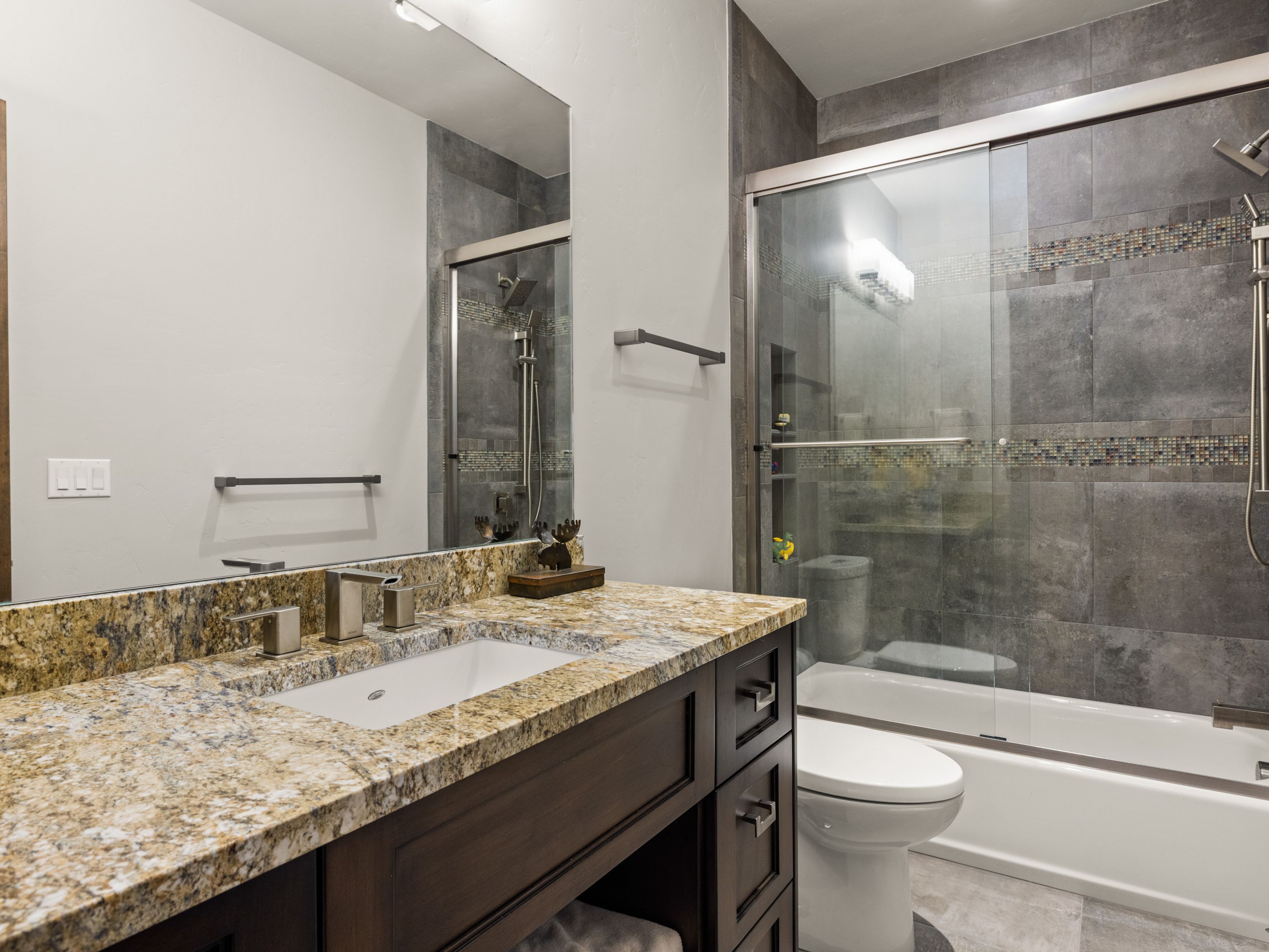 This bathroom features a dark wood vanity topped with a speckled stone countertop and an undermount sink. A glass-enclosed shower and tub combination is tiled with large gray tiles and a decorative mosaic accent strip. The space is completed by a white toilet and a large wall-mounted mirror, creating a functional and modern aesthetic.