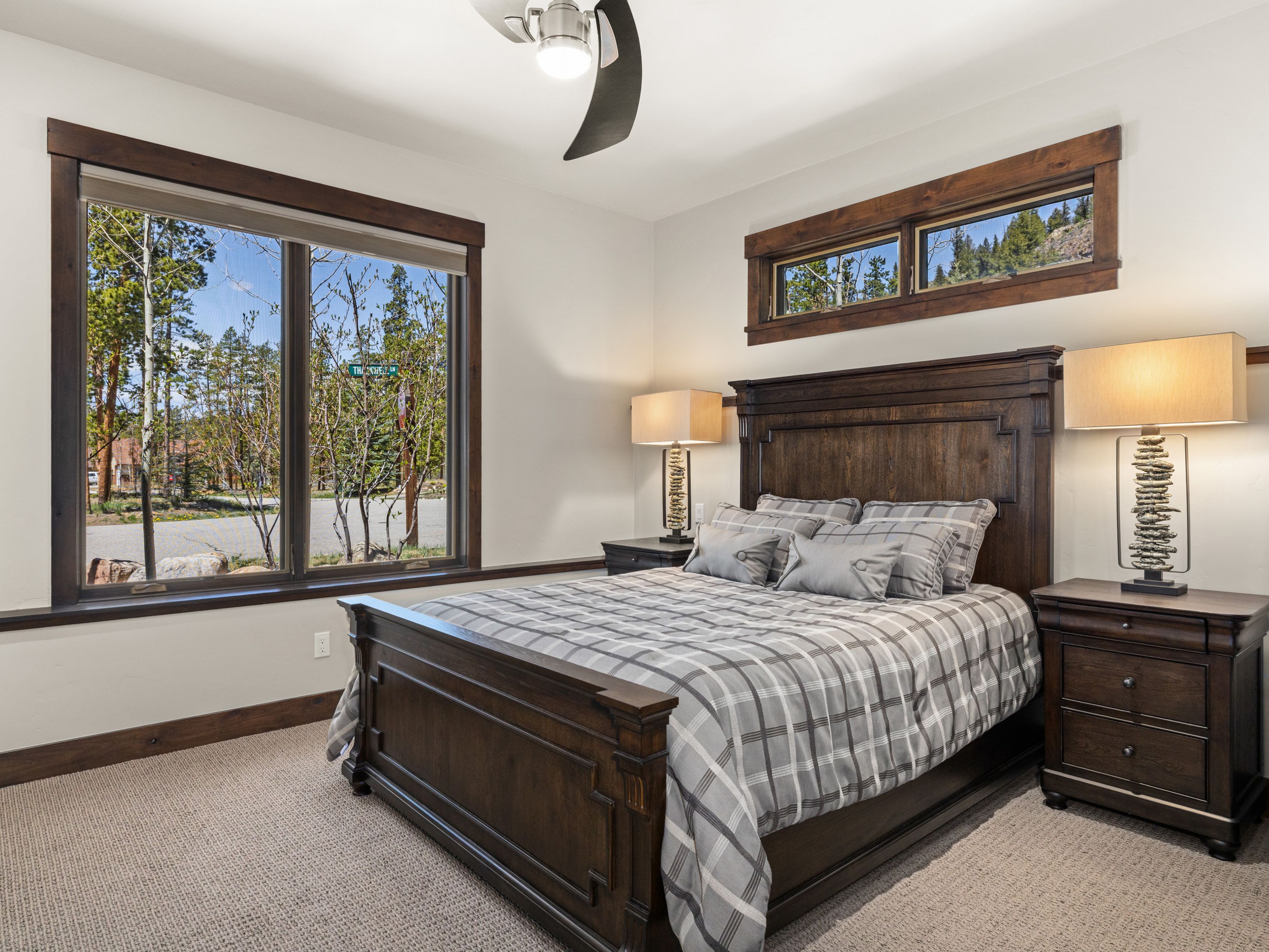 This bedroom features a dark wood bed frame with a matching headboard and nightstands, complemented by a gray plaid comforter. The room is accented by warm, textured table lamps and large windows with dark wood trim that frame a view of the surrounding trees. The neutral wall color and carpeted flooring create a cozy, inviting atmosphere suitable for a guest suite.