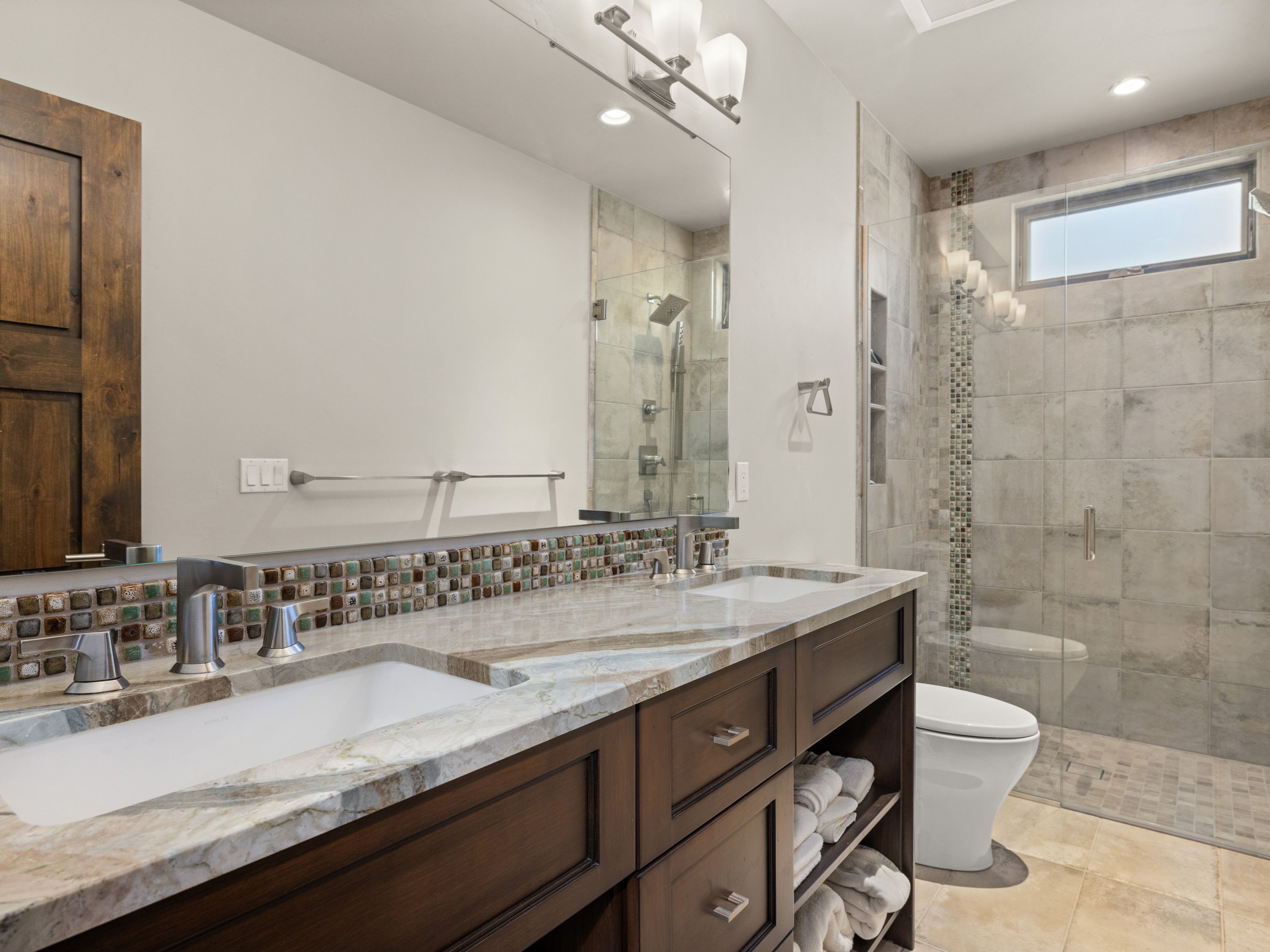 This primary bathroom features a double vanity with dark wood cabinetry and a patterned stone countertop. A mosaic tile backsplash adds texture, while a large mirror reflects the walk-in shower with glass doors and neutral-toned wall tiles. The space is well-lit by vanity lighting and a small window, creating a clean and functional atmosphere.