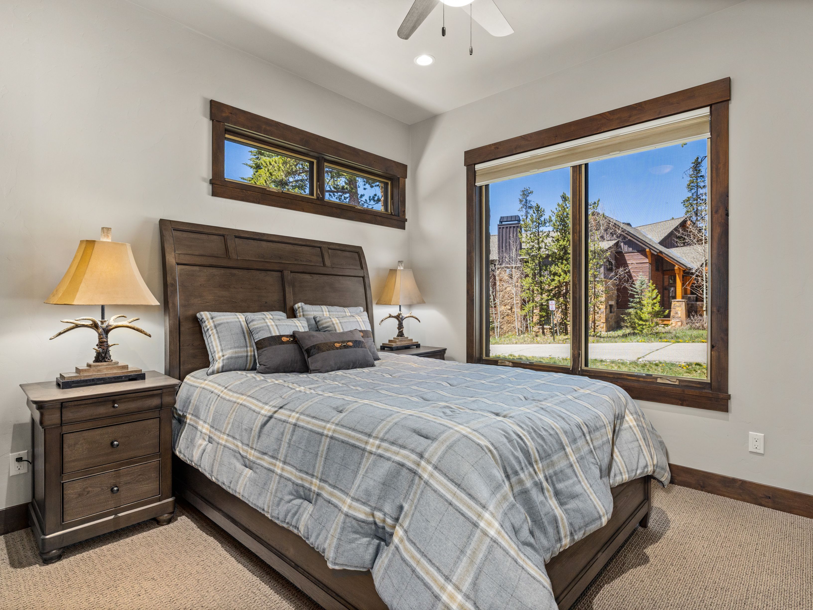 This cozy bedroom features a rustic aesthetic with dark wood furniture, including a substantial headboard and a matching nightstand topped with an antler-accented lamp. A plaid comforter in shades of blue and tan adds a classic, comfortable feel, while large windows provide ample natural light and a view of the surrounding wooded landscape. The room is finished with neutral walls and carpet, creating a warm and inviting atmosphere.
