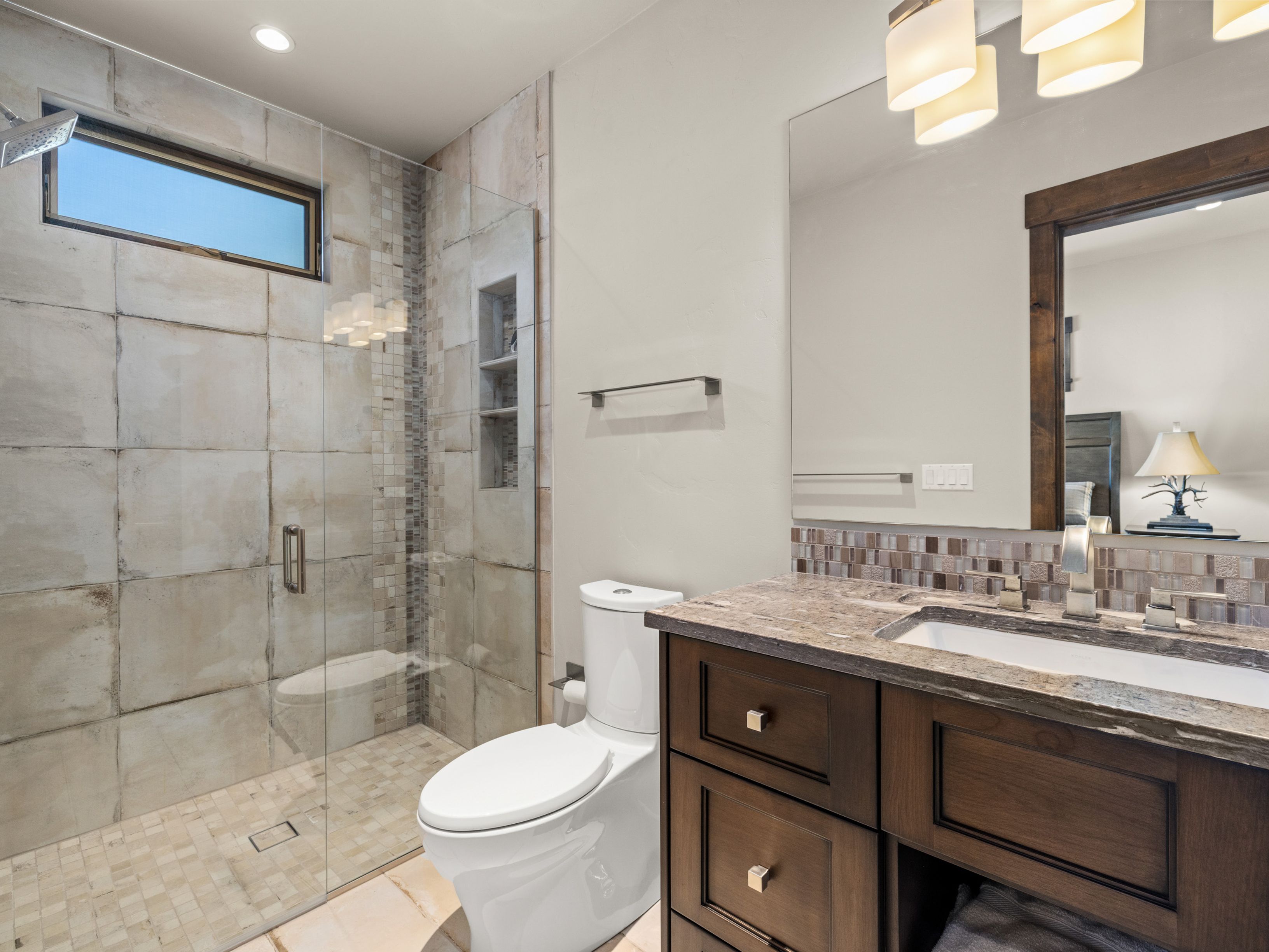 This modern bathroom features a dark wood vanity with a stone countertop and a rectangular undermount sink. To the left, a glass-enclosed walk-in shower is finished with large-format neutral tiles and a mosaic accent strip, complemented by a small transom window. The space is illuminated by a multi-light fixture above a large mirror, creating a clean and functional aesthetic.