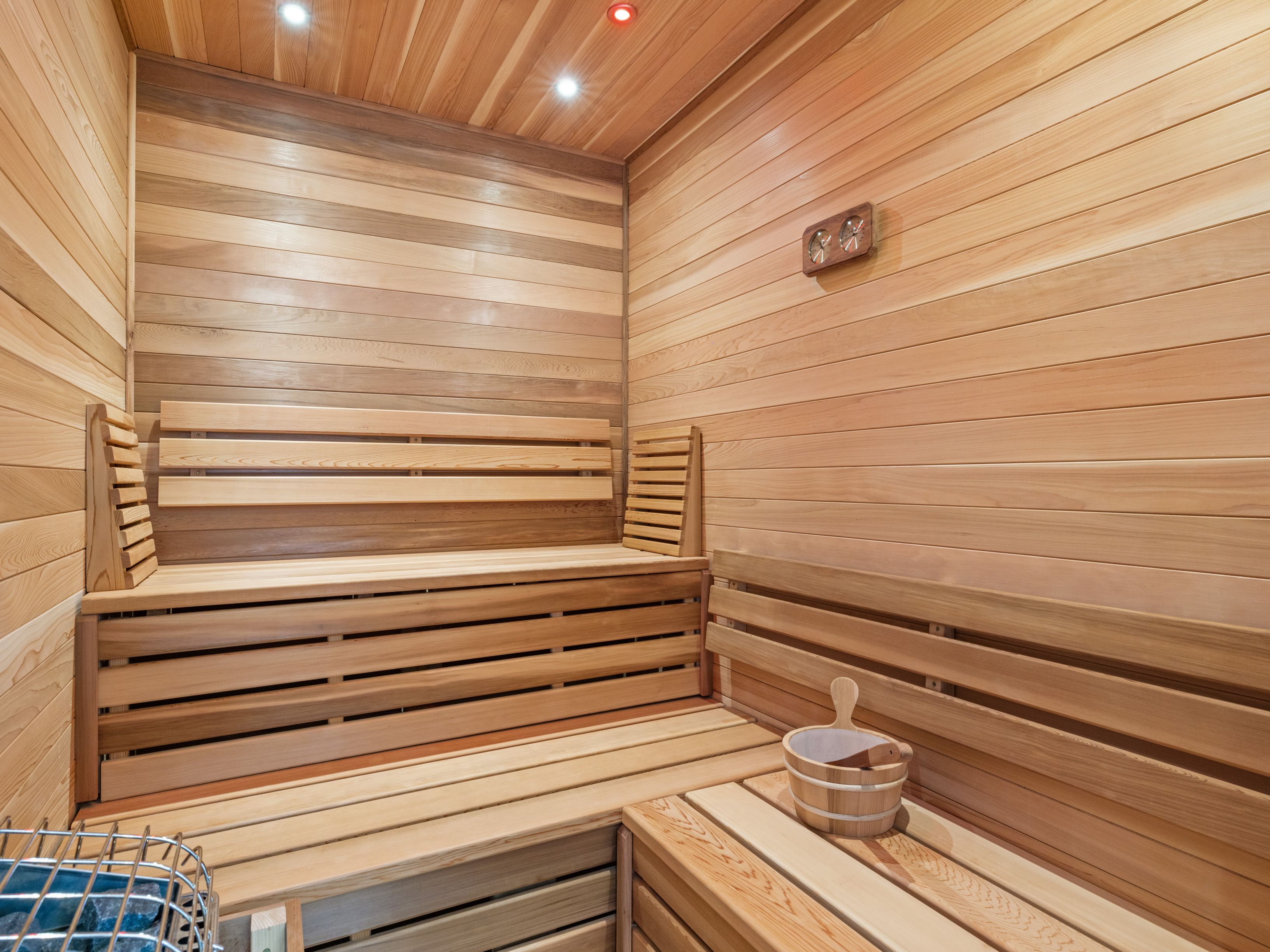 This interior view showcases a traditional sauna constructed with light-toned wood paneling on the walls and ceiling. The space features tiered wooden benches for seating, a wooden bucket with a ladle, and a wall-mounted thermometer, creating a warm and inviting atmosphere. The perspective is from inside the sauna, highlighting the clean, minimalist design and the integrated heating element in the foreground.