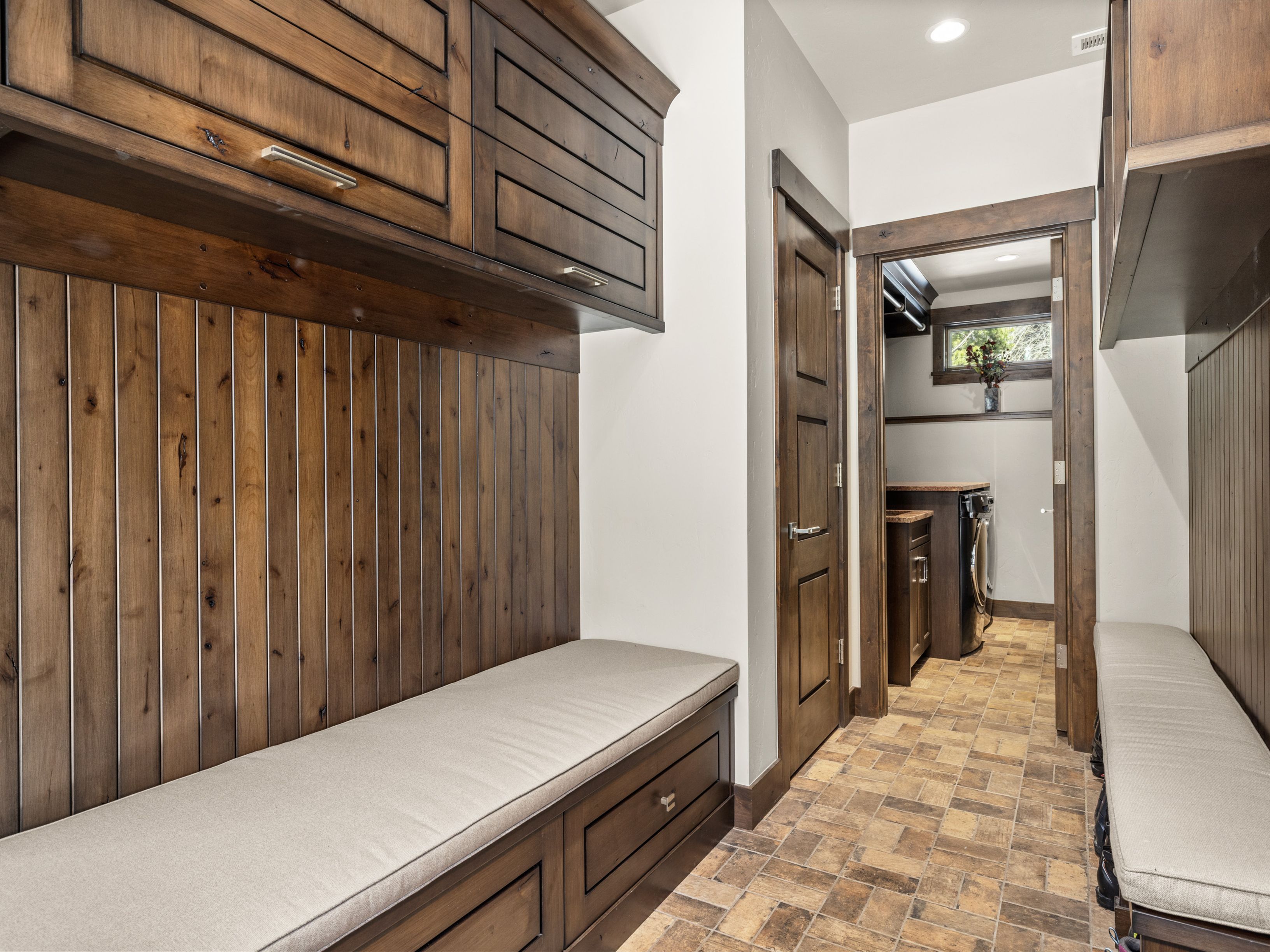 This mudroom area features custom built-in wooden cabinetry with integrated bench seating and light-colored cushions. The space is finished with vertical wood paneling and patterned tile flooring, leading through a doorway into a laundry room. The warm wood tones and rustic aesthetic create a functional and inviting transition space.