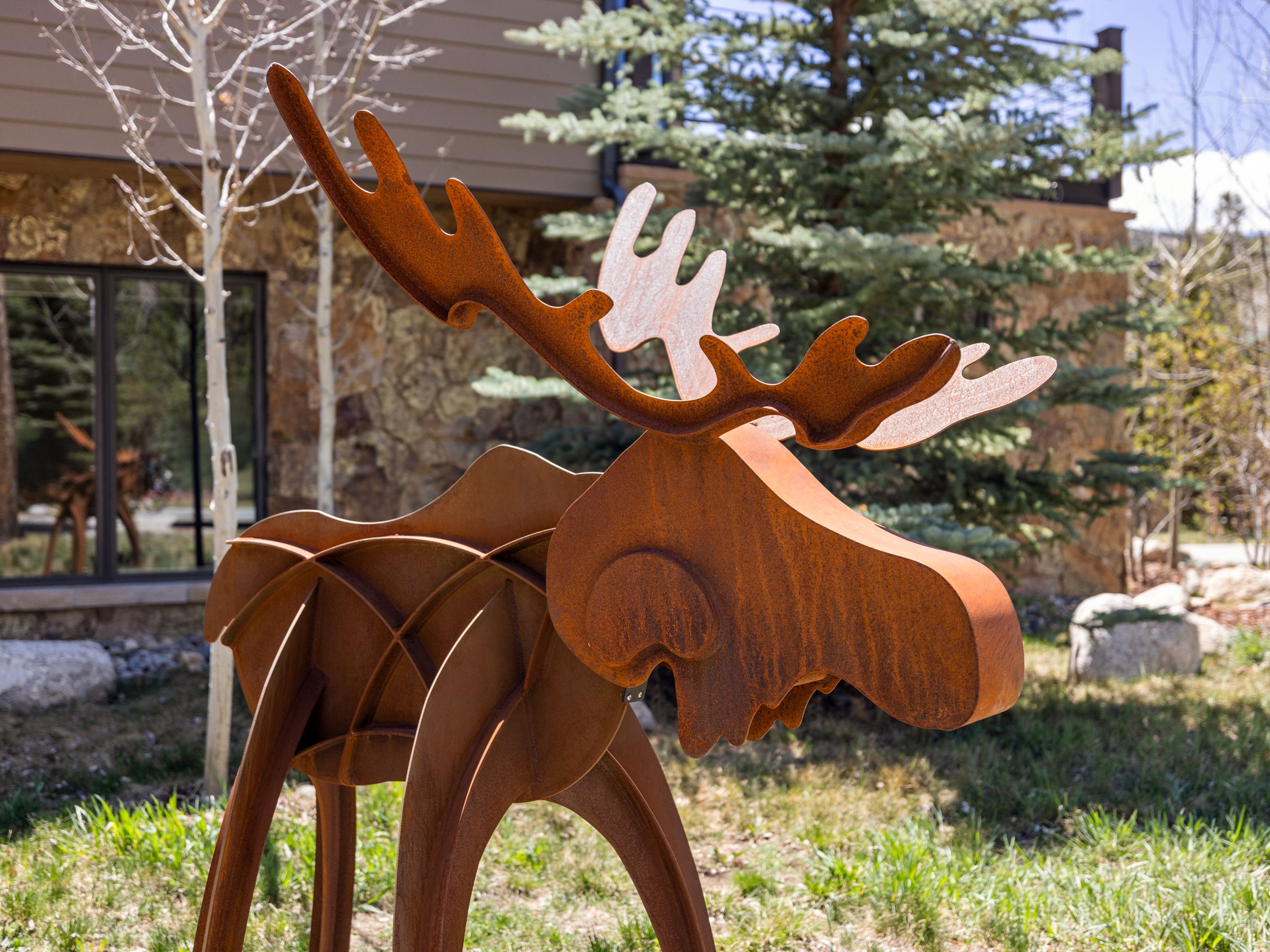 A sculptural, rust-toned metal moose figure stands prominently in a landscaped yard, serving as a unique focal point. The background features a stone-clad building exterior, mature evergreen trees, and a glimpse of a reflective window showing another sculpture. The scene captures a rustic, mountain-inspired aesthetic with natural lighting highlighting the textured metal surface.