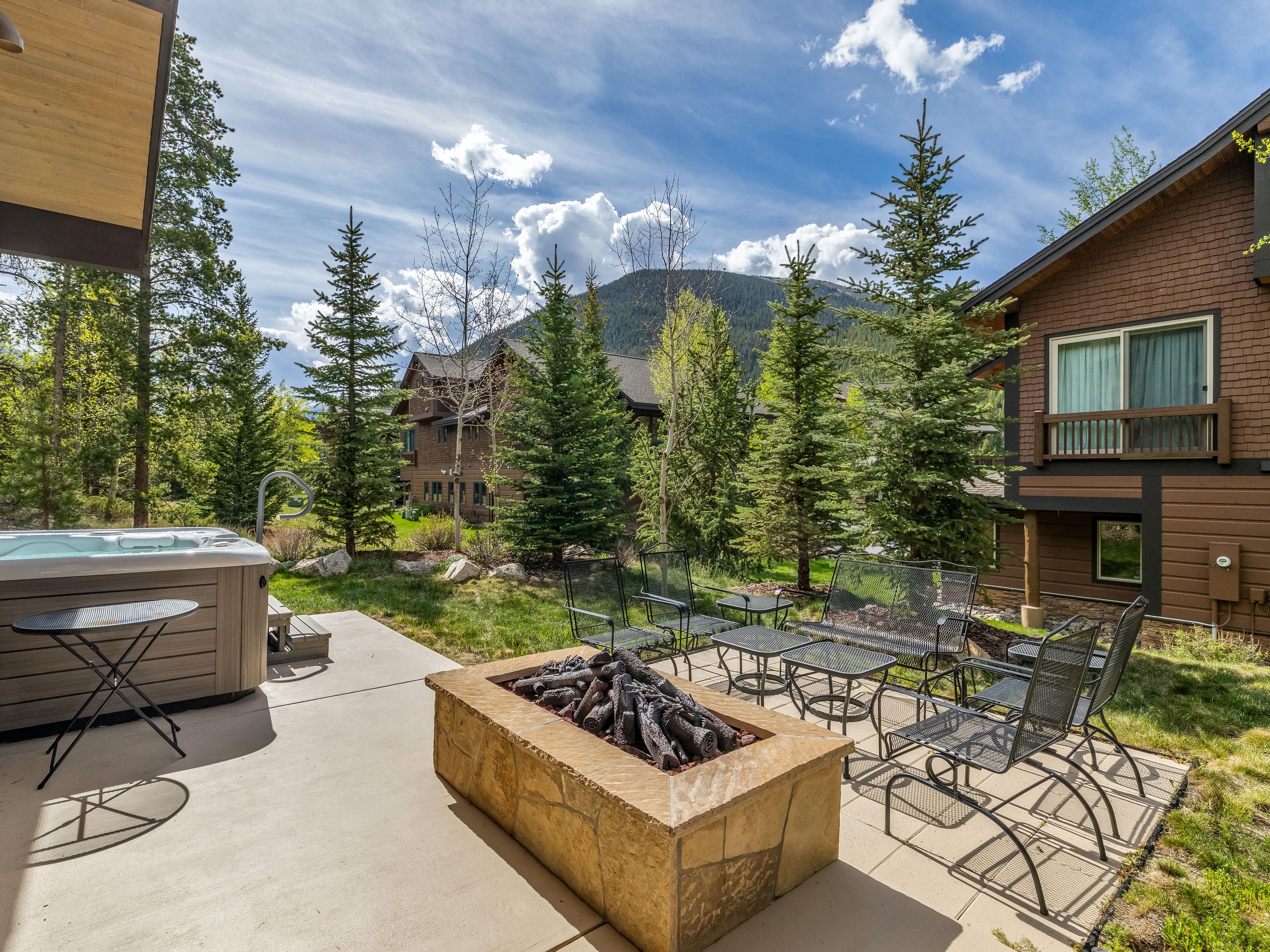 This inviting outdoor patio space features a stone-clad fire pit as the central focal point, surrounded by a set of metal mesh patio chairs and tables. To the left, a hot tub sits on the concrete slab, while the surrounding landscape includes lush green grass and tall pine trees against a backdrop of mountain peaks. The scene captures a serene, mountain-resort atmosphere, perfect for relaxation and outdoor entertaining.