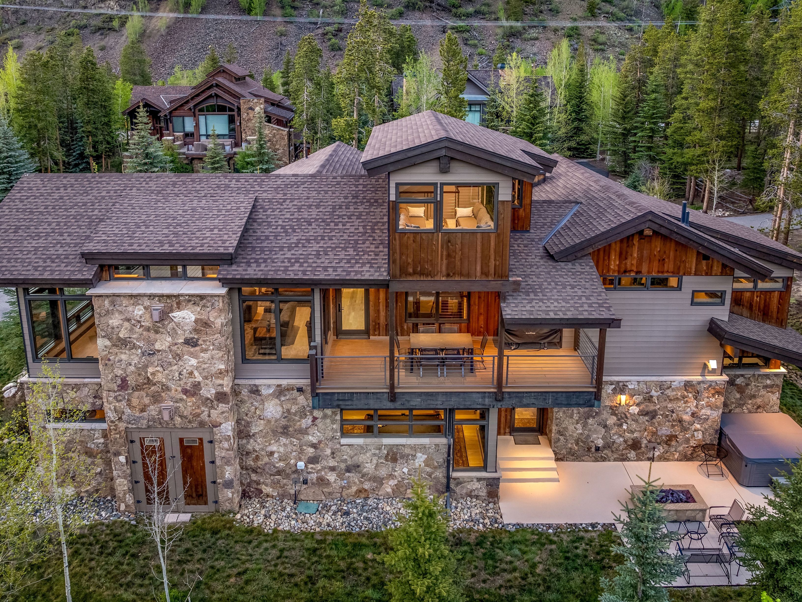 This elevated aerial perspective captures a luxurious mountain residence featuring a blend of natural stone and warm wood siding. The home includes a spacious elevated deck with outdoor dining, a lower-level stone patio with a fire pit, and a private hot tub area. Surrounded by lush evergreen trees and a rugged hillside, the property exudes a sophisticated, rustic aesthetic perfect for a mountain retreat.