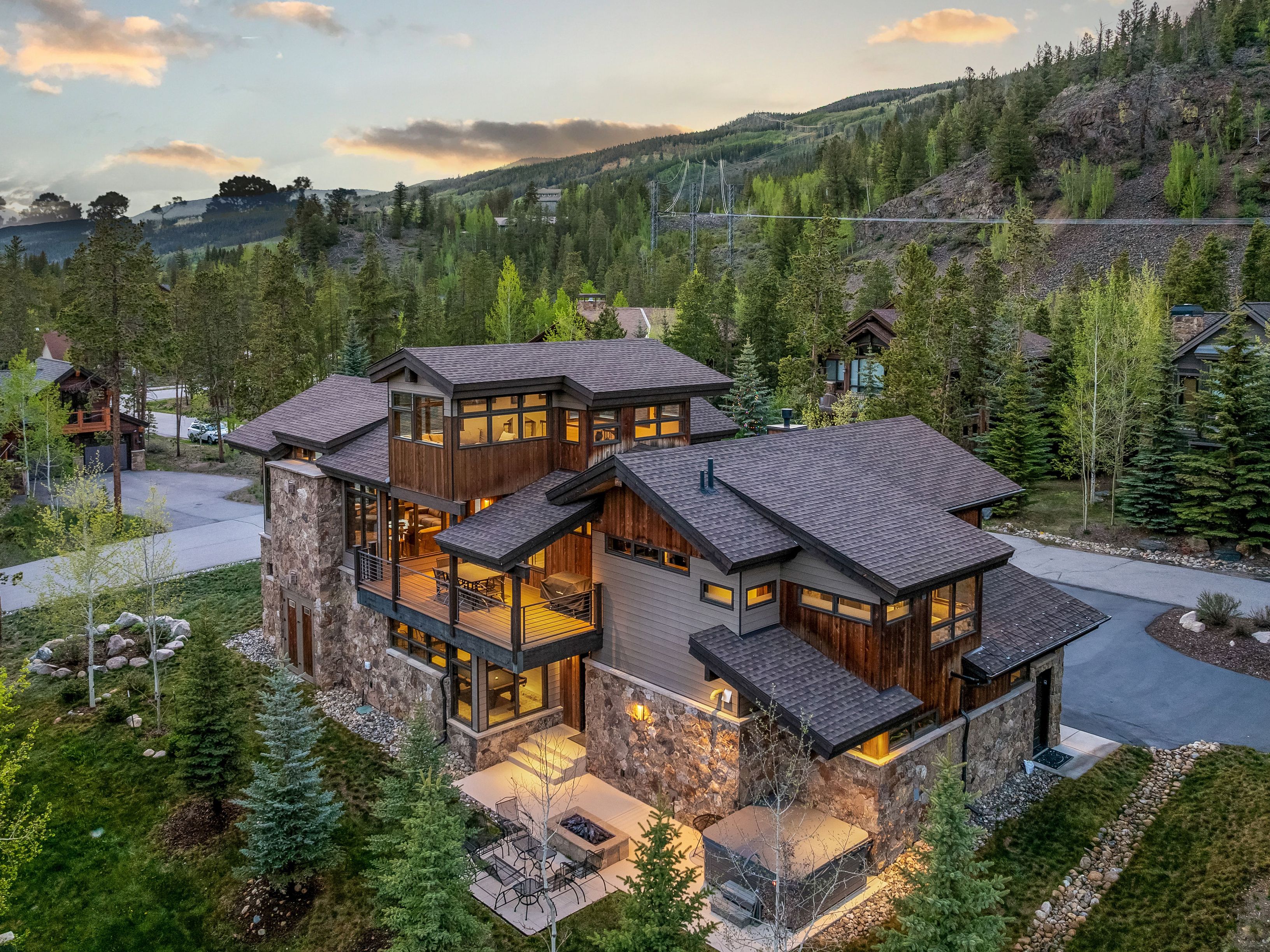 This high-angle aerial shot captures a luxurious mountain residence characterized by a blend of rustic stone masonry and warm wood siding. The multi-level structure features expansive decks, a private patio with a fire pit, and a hot tub, all nestled within a lush, forested landscape. The architectural design emphasizes a modern mountain aesthetic, perfectly integrated into the sloping terrain under a soft, golden-hour sky.