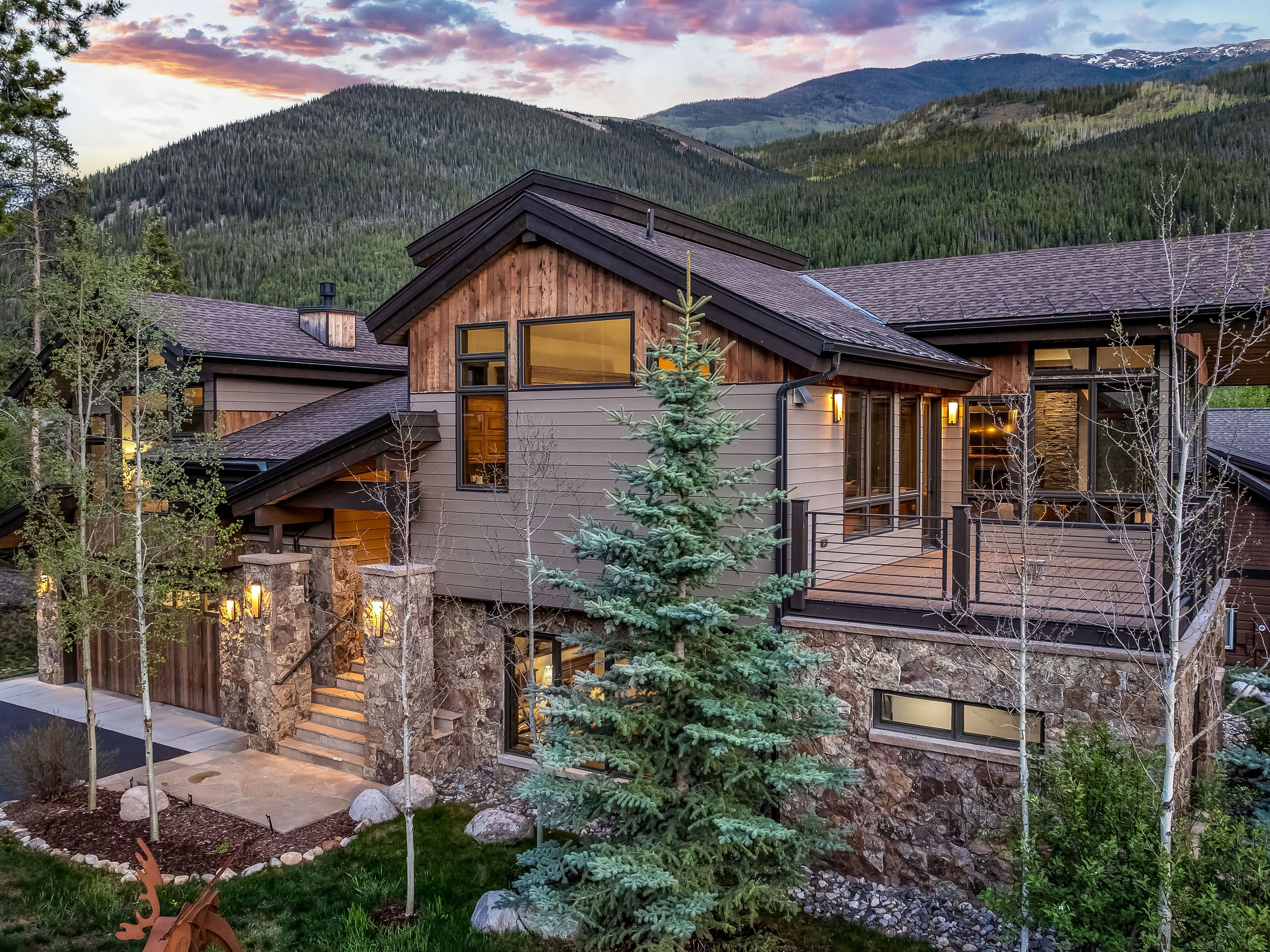 This stunning mountain residence showcases a sophisticated blend of rustic and modern architectural styles, featuring a combination of natural stone masonry, horizontal siding, and warm wood accents. The home is nestled against a backdrop of lush, forested mountains under a dramatic sunset sky, with a prominent elevated deck and a welcoming stone-stepped entryway. Large windows and warm exterior lighting create an inviting atmosphere, highlighting the home's integration into its rugged, scenic environment.