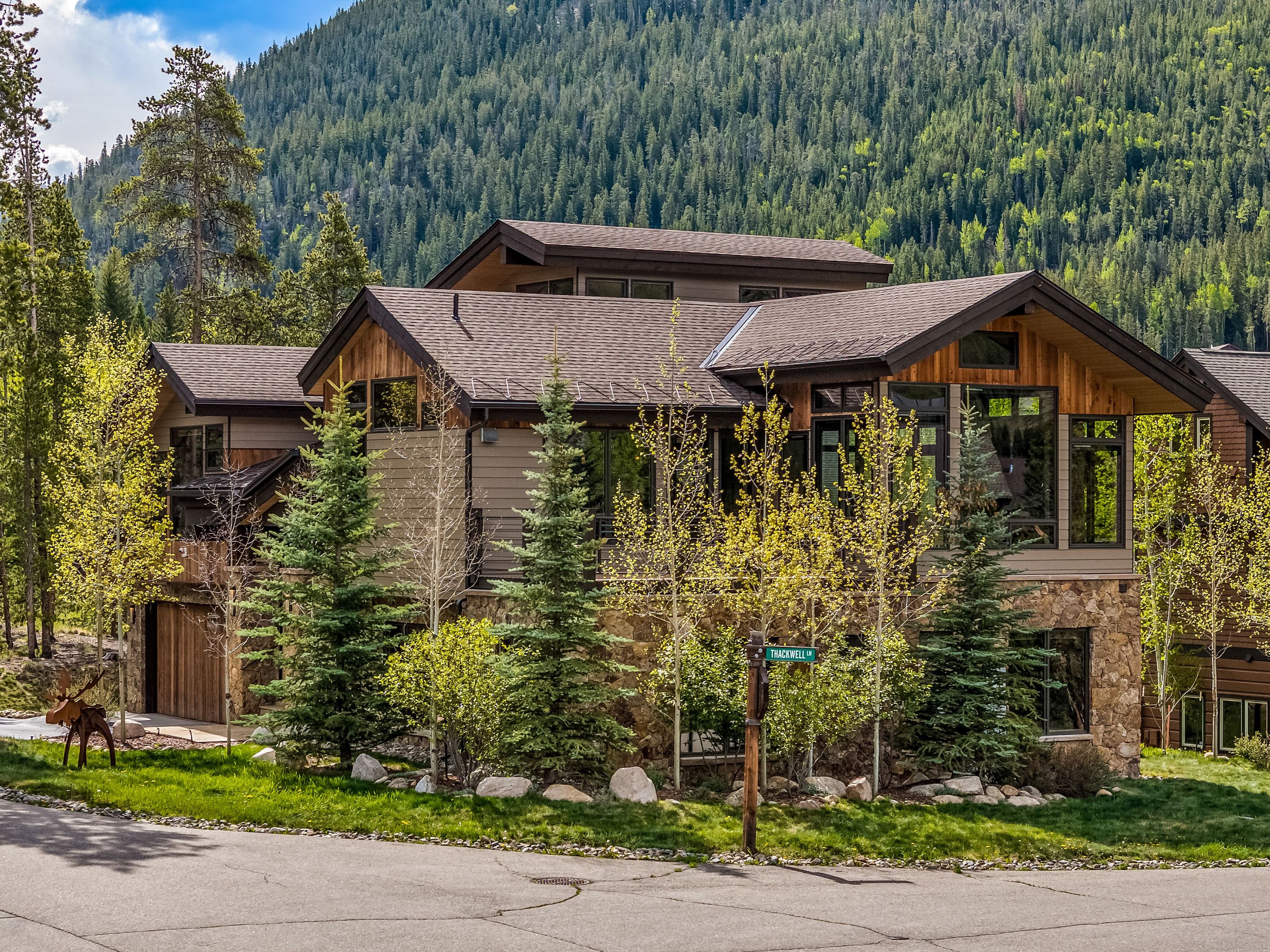 This mountain-style residence features a multi-gabled roofline, a mix of horizontal siding and natural wood accents, and a stone-clad base. Large windows provide expansive views of the surrounding forest and mountain backdrop, while a metal moose sculpture stands on the front lawn near a street sign. The home is nestled in a lush, wooded landscape, creating a serene and upscale retreat atmosphere.