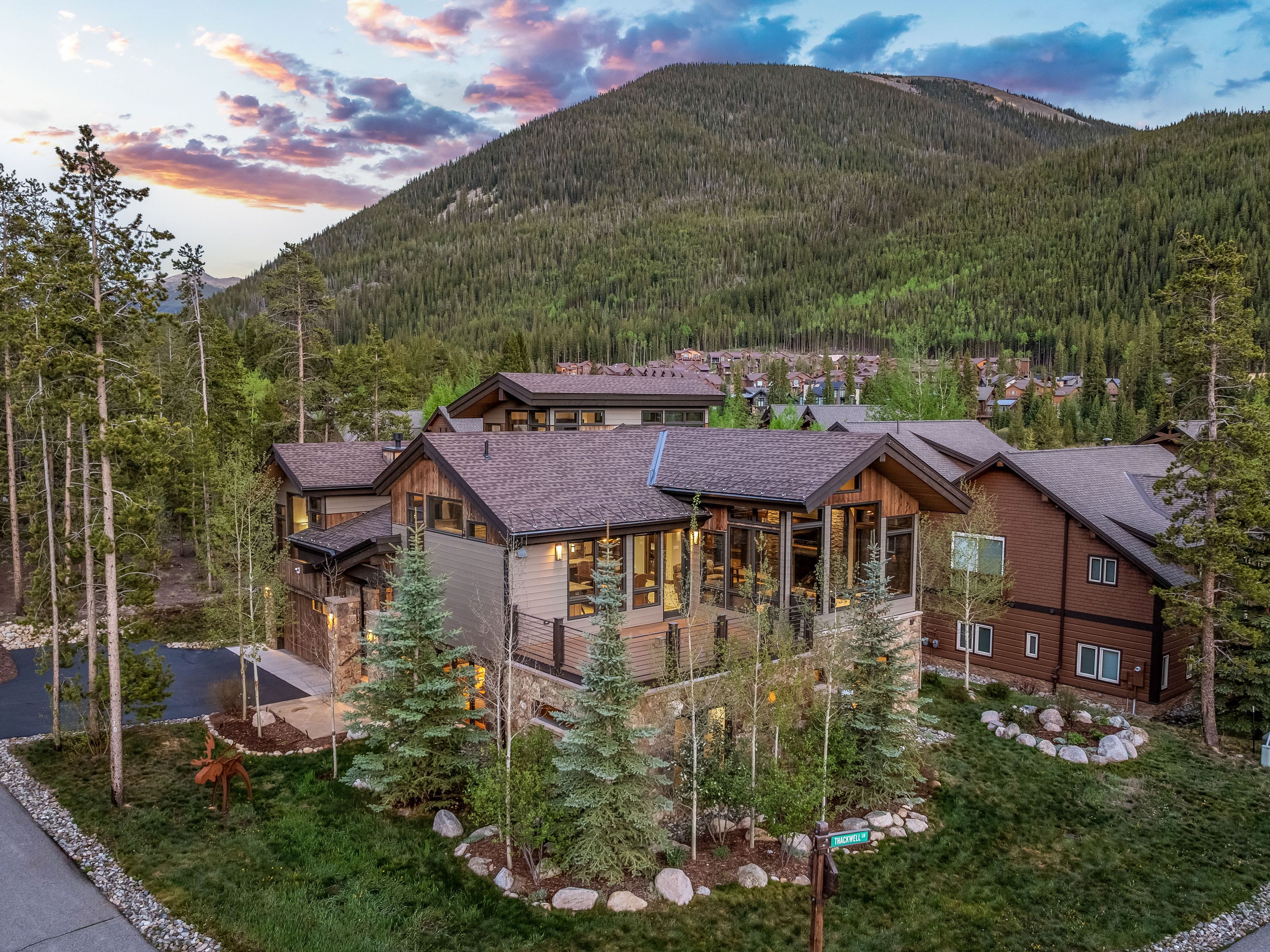 This aerial perspective captures a modern mountain home nestled in a lush, forested landscape at dusk. The structure features a blend of stone and wood siding, with a prominent elevated deck and large windows that reflect the soft evening light. The surrounding environment includes dense pine trees and a backdrop of a large mountain, creating a serene and upscale residential atmosphere.