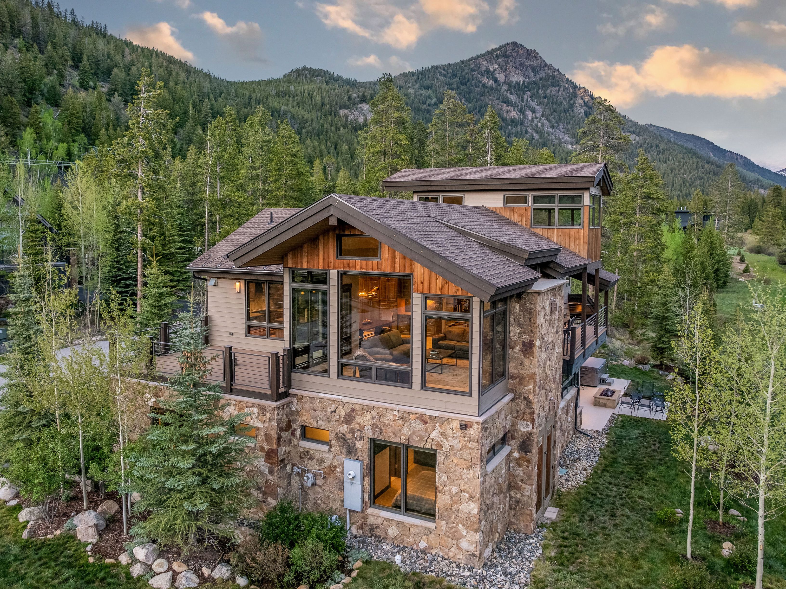 This aerial perspective captures a modern mountain home nestled within a lush, forested landscape. The structure features a combination of stone masonry on the lower level and warm-toned wood siding on the upper levels, complemented by large windows that offer expansive views of the surrounding pines and distant peaks. A spacious deck extends from the main living area, while a stone patio with a fire pit sits at the base, creating a seamless transition between the home and its natural environment.