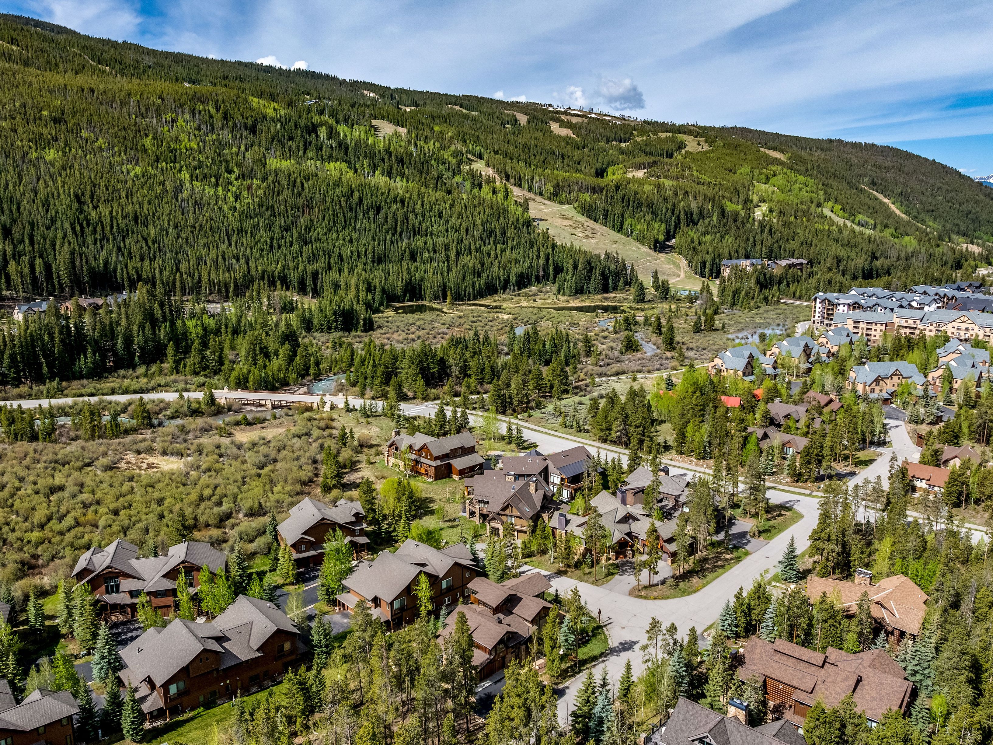This high-angle aerial view captures a mountain resort community nestled at the base of a lush, forested ski slope. The scene features a collection of multi-story, wood-sided homes with pitched roofs, integrated into a landscape of pine trees and winding paved roads. A bridge spans a small stream in the foreground, leading toward a larger complex of buildings in the distance, creating a serene and picturesque alpine atmosphere.