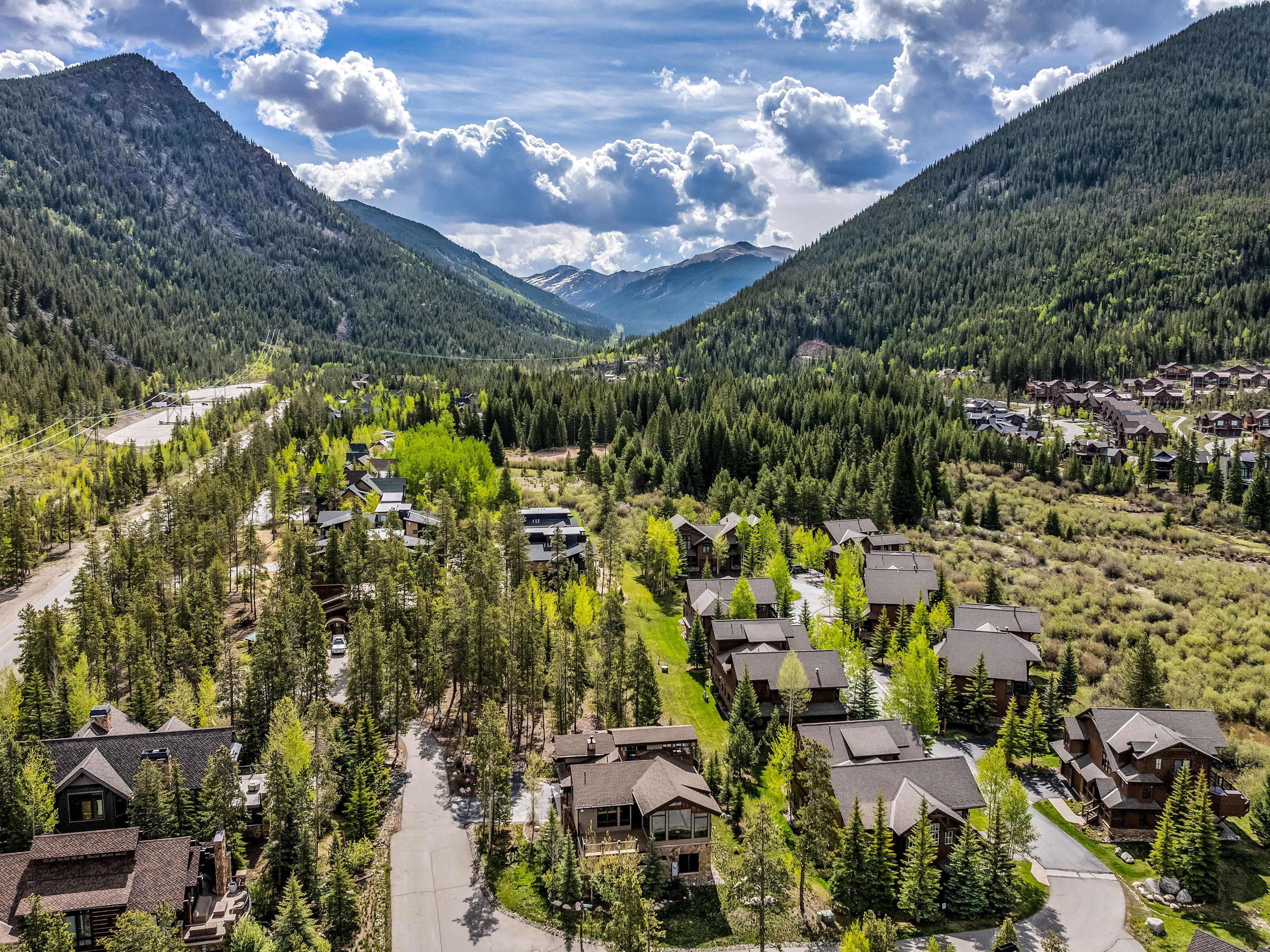 This high-angle aerial view captures a mountain residential community nestled within a lush, forested valley surrounded by towering, pine-covered peaks. The scene features multiple multi-story homes with dark-toned roofs and wood-accented exteriors, connected by winding paved roads and interspersed with vibrant green trees. The perspective provides a cinematic sense of scale, highlighting the serene, secluded nature of the neighborhood against the backdrop of a dramatic, cloud-filled sky.