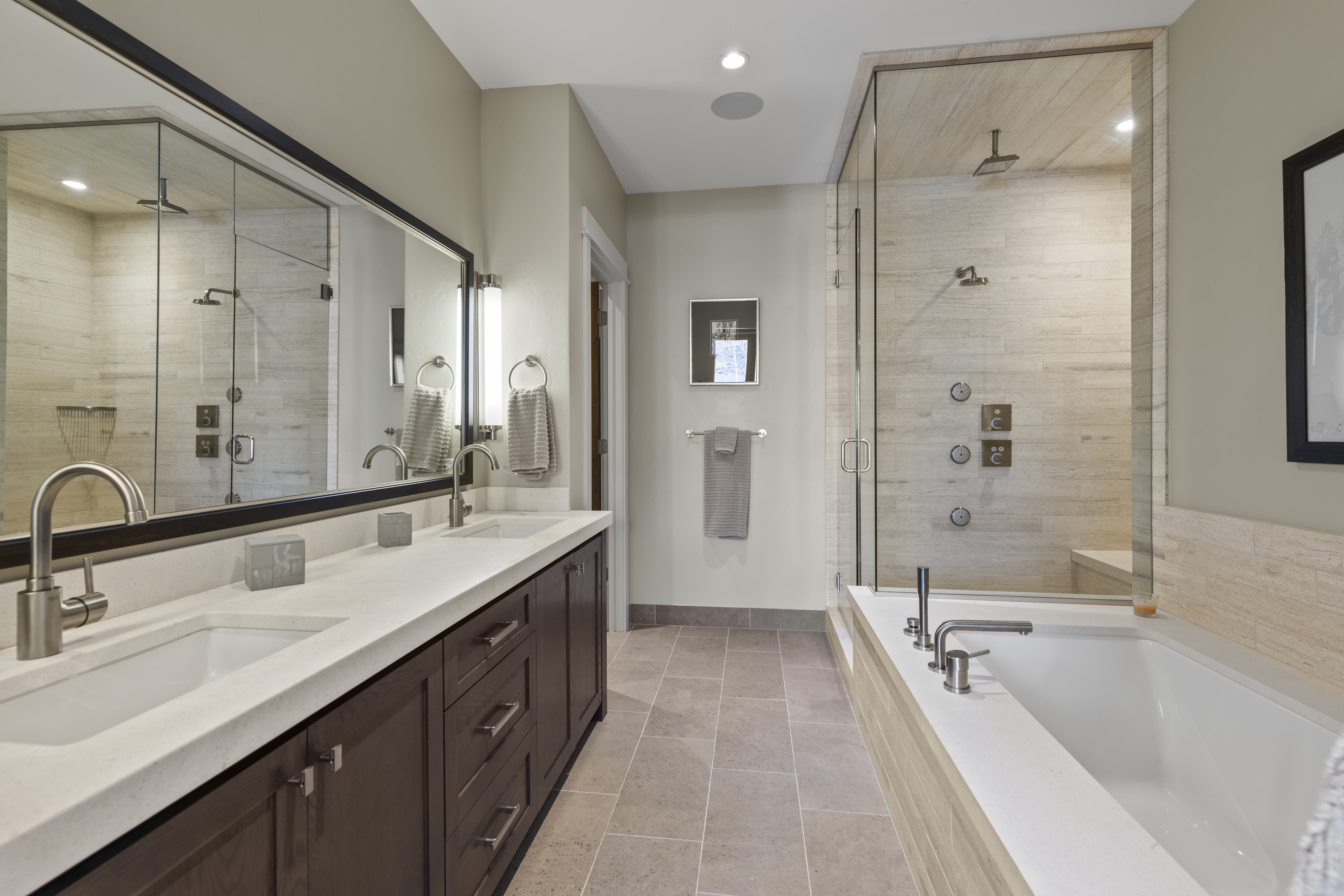 This is a well-lit primary bathroom featuring a double vanity with dark wood cabinets and a light countertop. A large mirror with a dark frame hangs above the sinks, reflecting the adjacent shower area. The shower and tub are enclosed with glass and feature modern fixtures, creating a spa-like atmosphere.