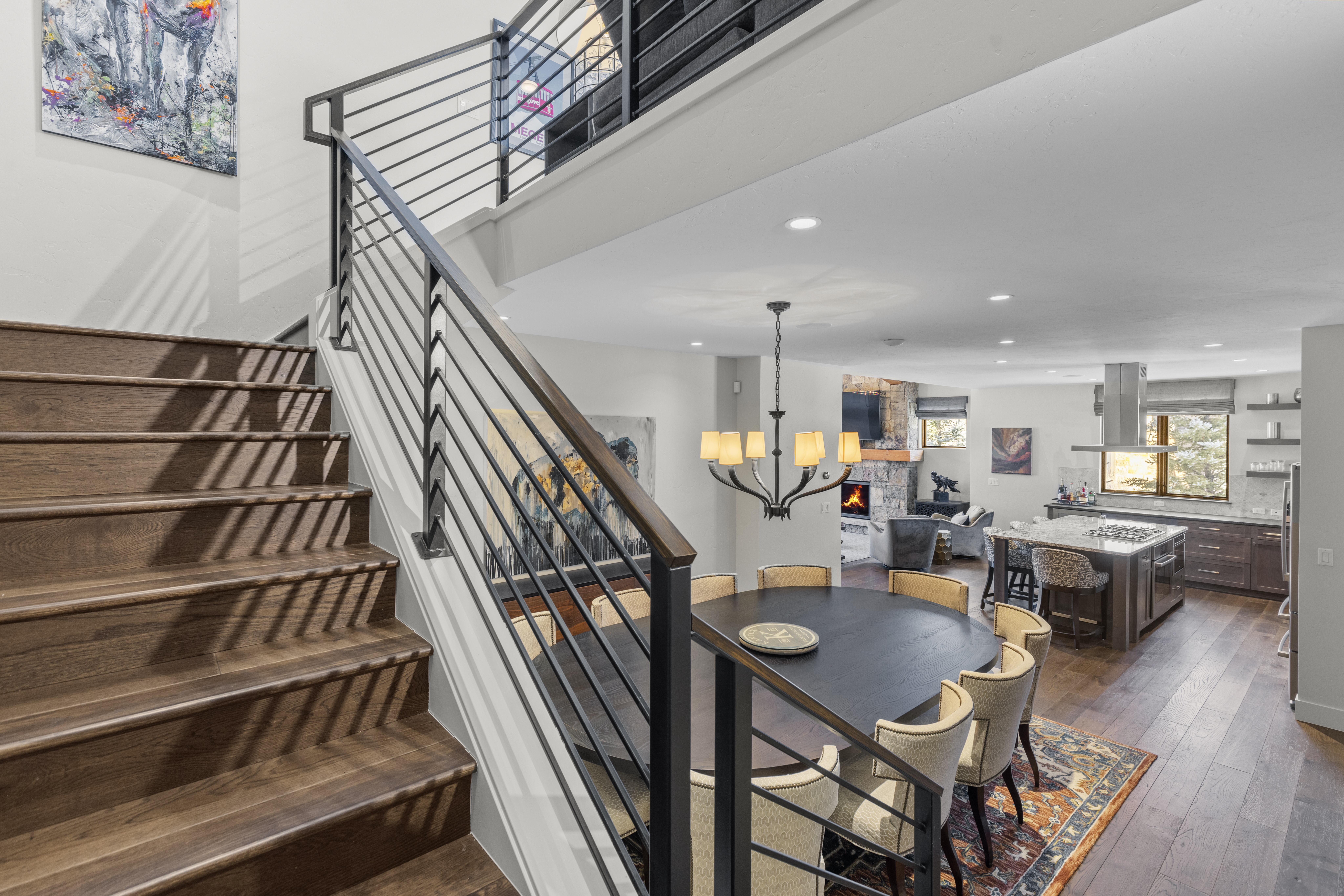 This interior shot showcases a dining area adjacent to a modern kitchen and living space. A dark, round dining table is surrounded by upholstered chairs, illuminated by a chandelier. The space is connected by hardwood flooring and features a staircase with metal railings, creating an open and inviting atmosphere.