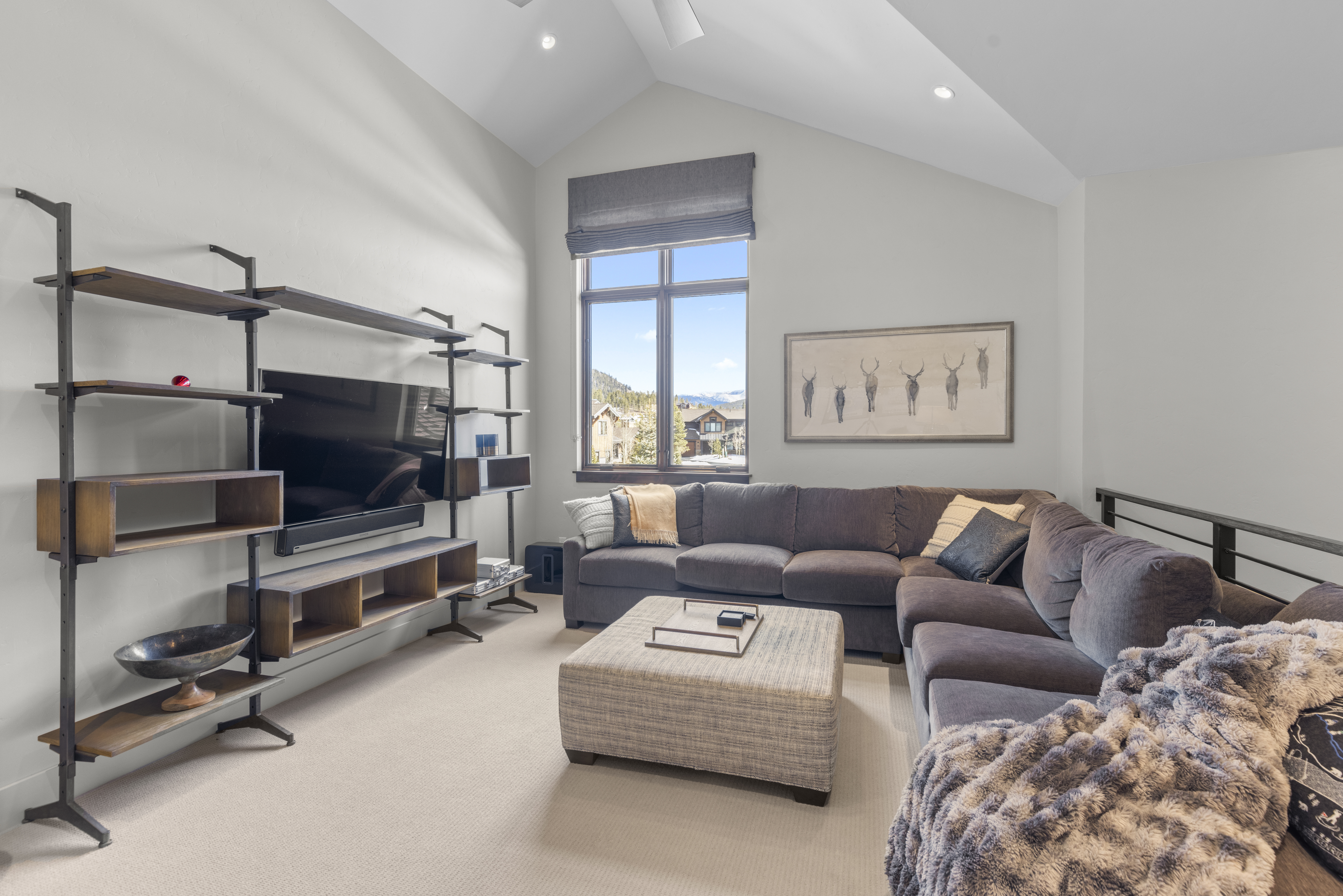 This is an interior shot of a living room featuring a large, sectional sofa in a dark gray color, complemented by a textured ottoman. A modern shelving unit with a television is positioned against one wall, while a window with a gray shade offers a view of the outside. The room is well-lit and appears cozy and inviting, with neutral-toned carpet and artwork adding to the aesthetic.