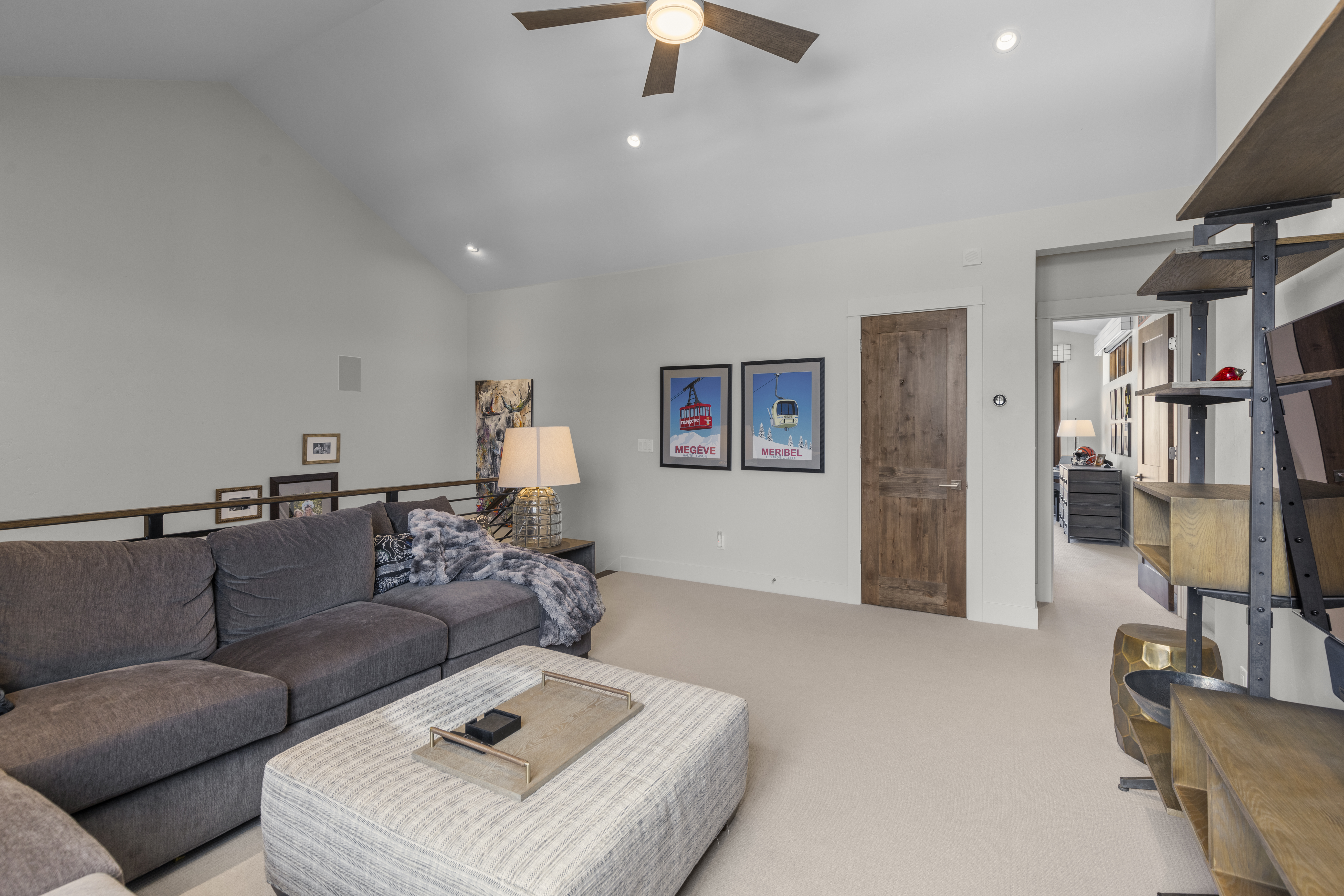 This is an interior shot of a living room featuring a large gray sectional sofa, a striped ottoman, and a wooden door. Two framed posters hang on the wall, and a unique shelving unit stands to the right of the door. The room is well-lit with a ceiling fan and recessed lighting, creating a comfortable and inviting atmosphere.
