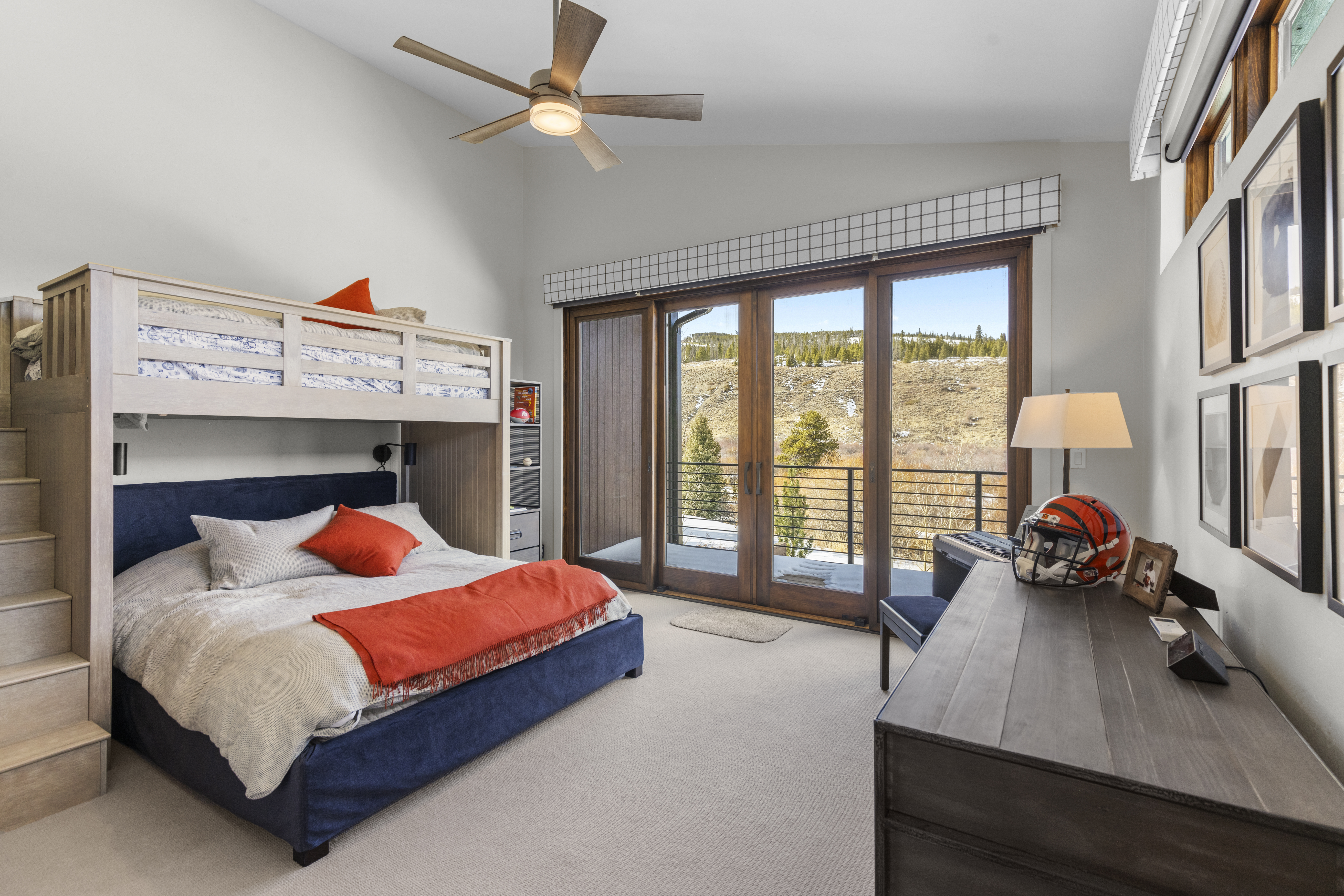 This is a well-lit bedroom featuring a bunk bed with a blue lower bed and a wooden frame, complemented by orange pillows and a throw. A wooden desk with a lamp and decorative items sits near a sliding glass door that leads to a balcony with a view of a hillside. The room has a neutral color palette with carpet flooring, creating a cozy and inviting atmosphere.
