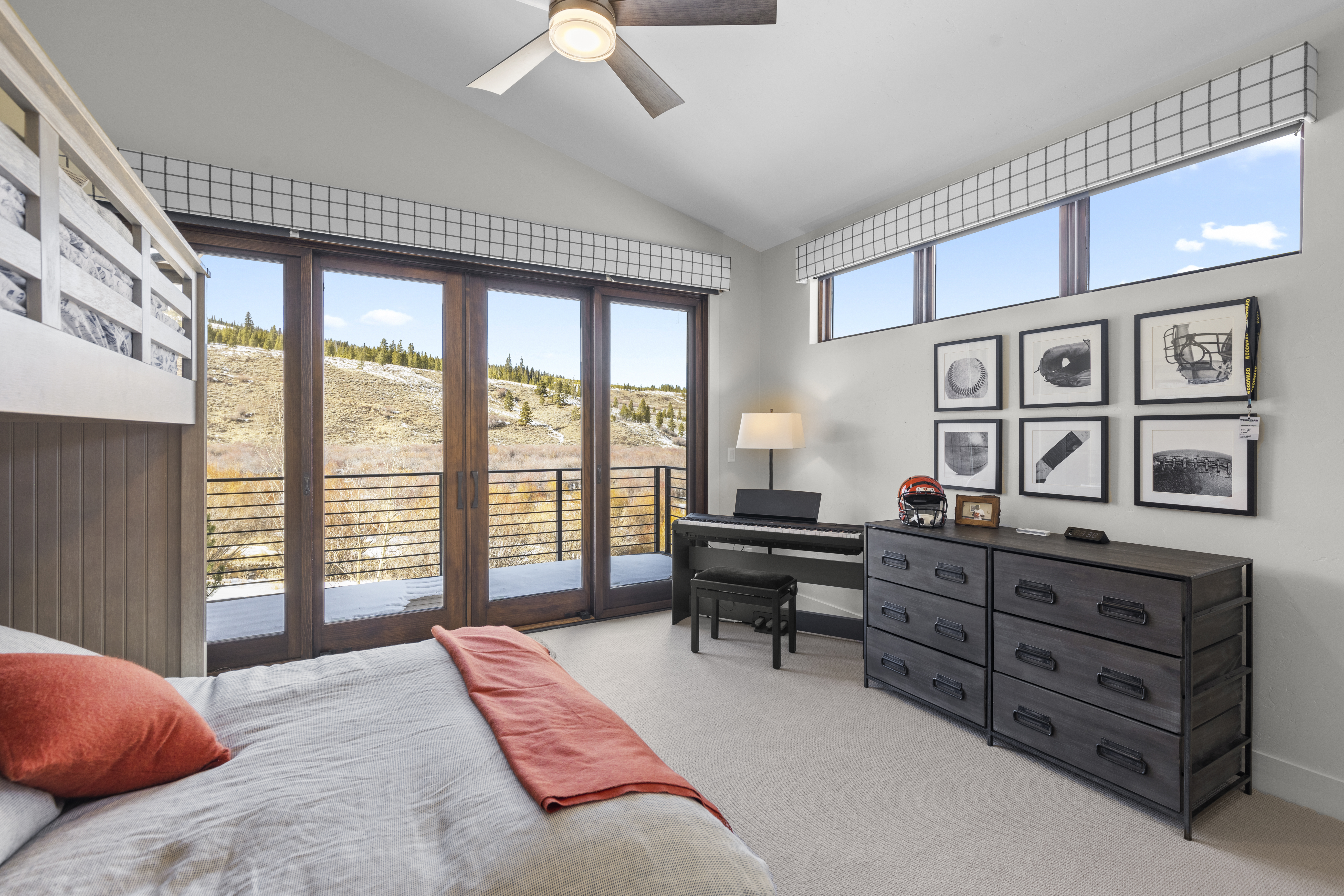This is a well-lit bedroom featuring a bed with orange accents, a dark wood dresser with sports memorabilia, and a black piano with a bench. Large sliding glass doors lead to a balcony with a view of a hillside, and a row of framed sports-themed artwork hangs above the dresser. The room has a modern and comfortable feel.