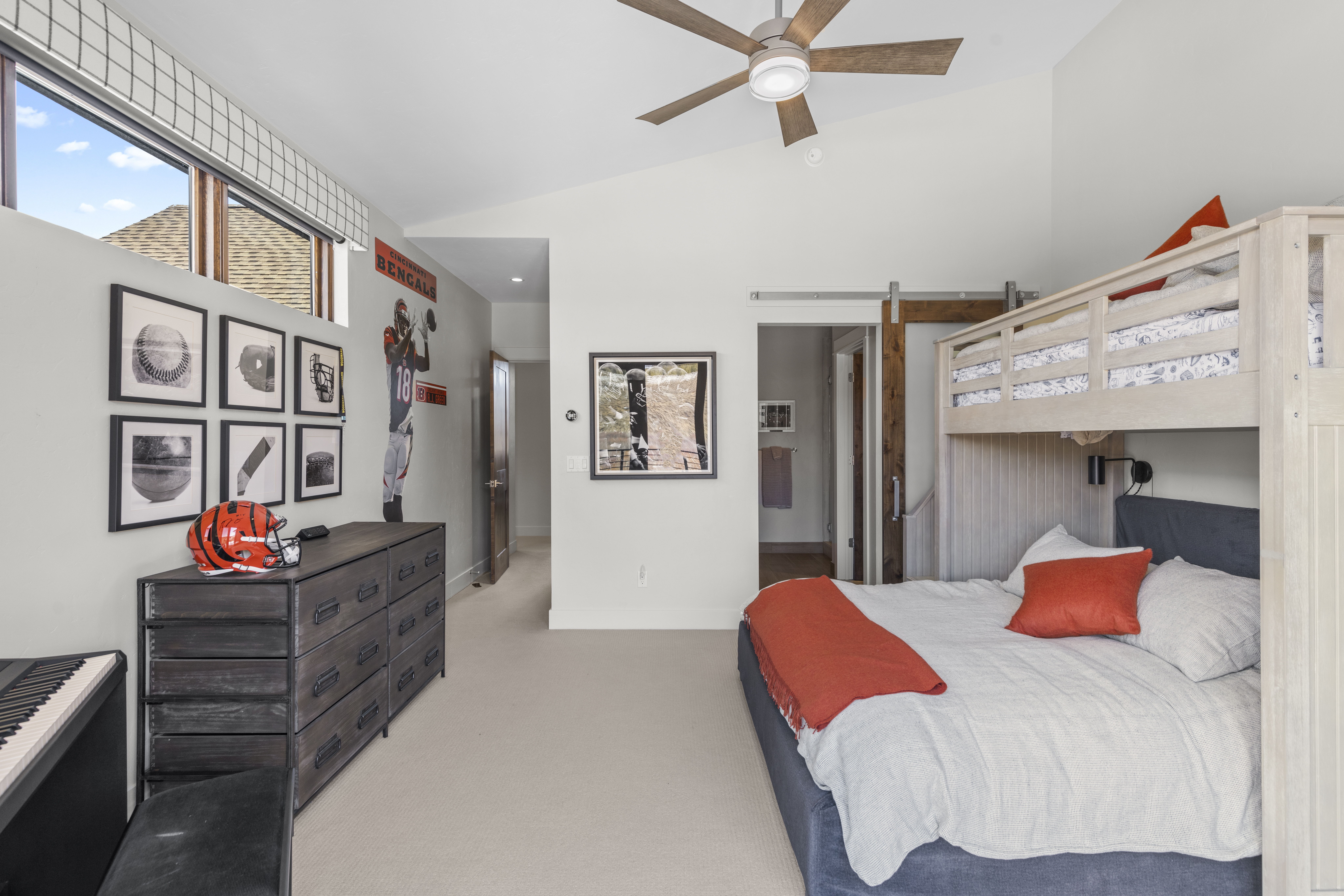 This bedroom features a bunk bed with a full-sized bed below, complemented by an orange throw and pillow. A dark wood dresser sits against the wall, adorned with sports memorabilia, and framed artwork adds a personal touch. The room is well-lit and has a cozy, inviting atmosphere.