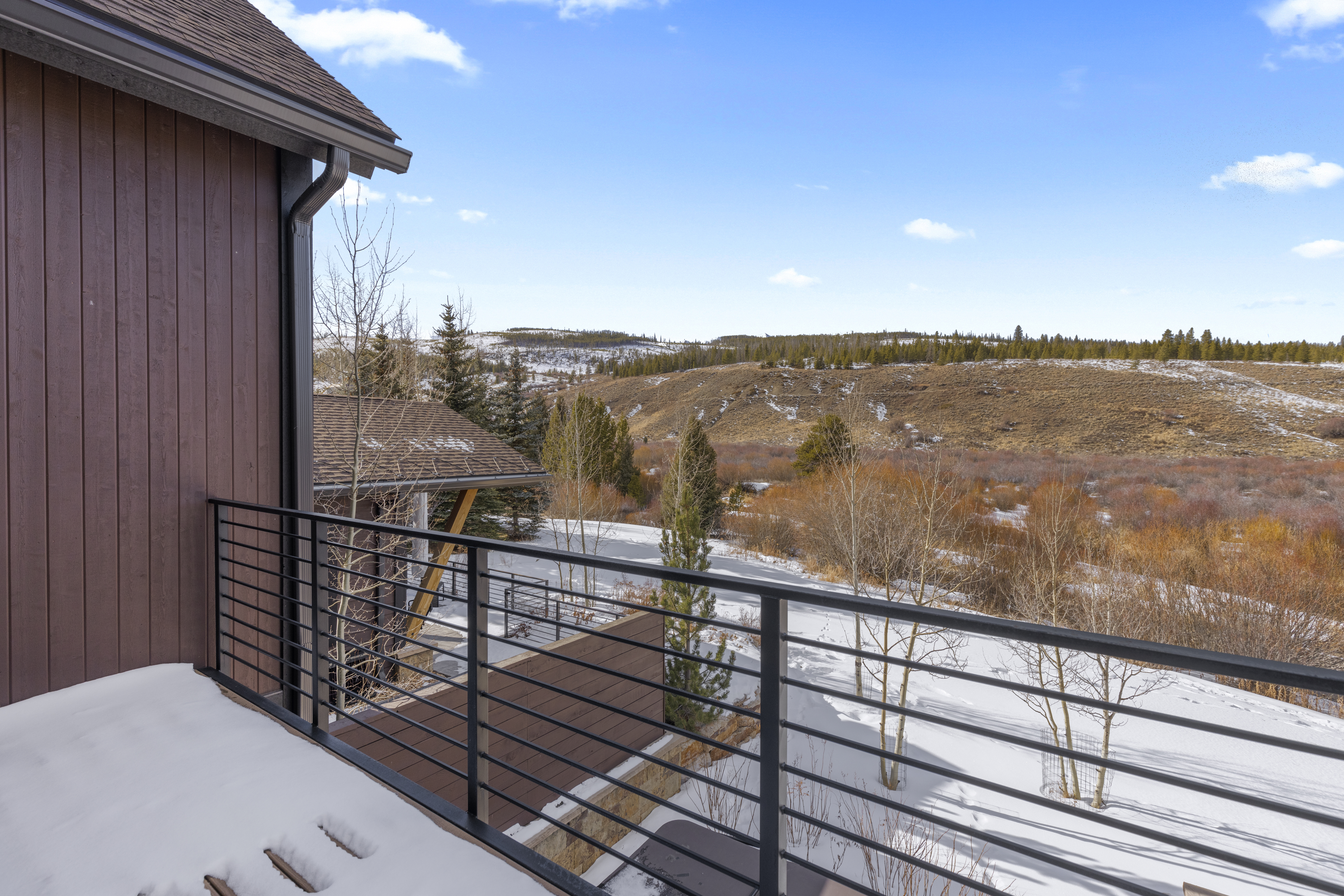 This image showcases a balcony with a modern horizontal railing, offering a scenic view of a snow-covered landscape with rolling hills and scattered trees. The balcony features a dark-stained deck surface, and the exterior of the adjacent building is clad in brown siding. The overall impression is one of tranquility and connection to nature.