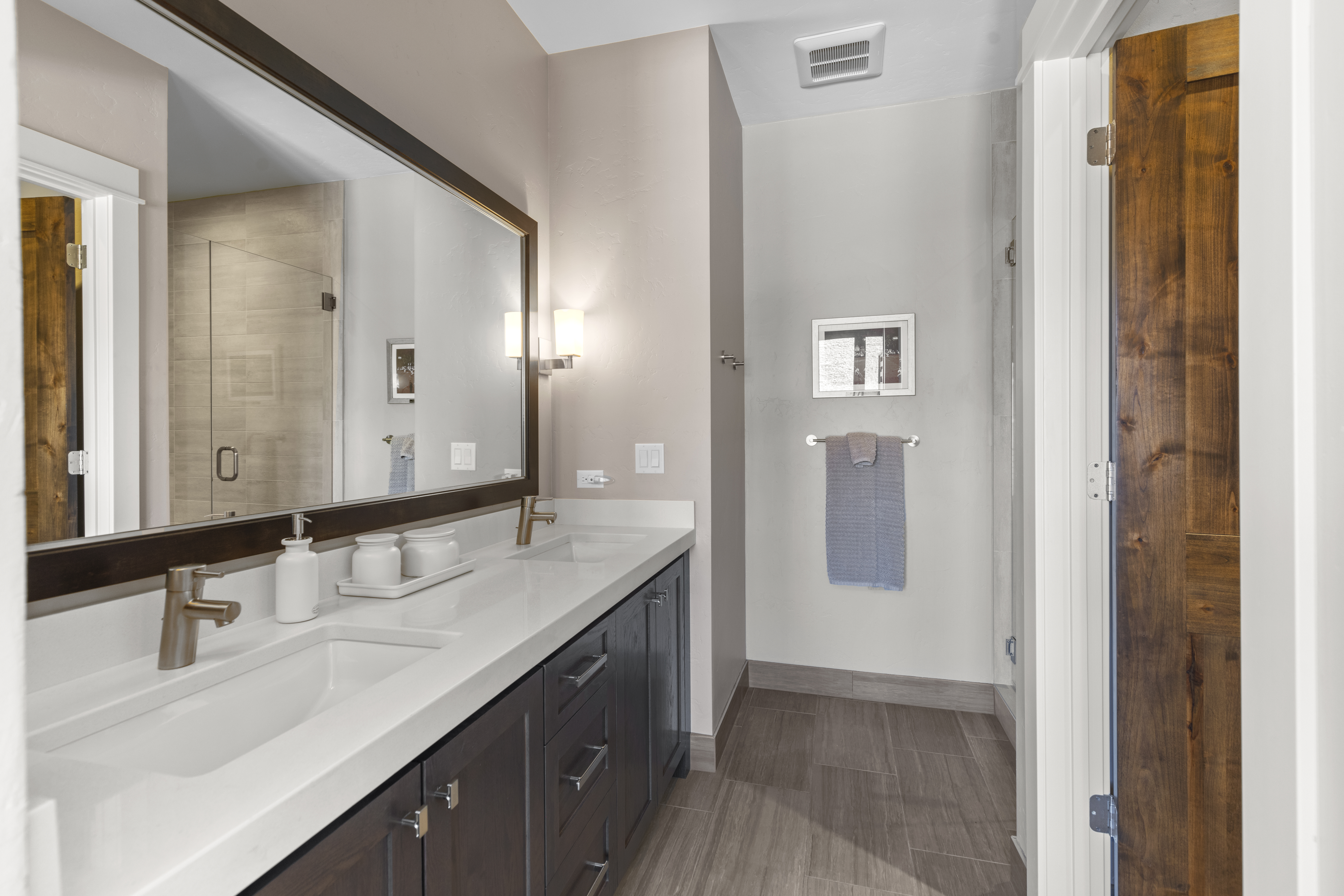 This is a well-lit primary bathroom featuring a double vanity with white countertops and dark wood cabinetry. A large mirror hangs above the sinks, reflecting a glimpse of the shower. The room has a modern aesthetic with neutral wall colors and wood-look tile flooring, leading to a partially visible wooden door.