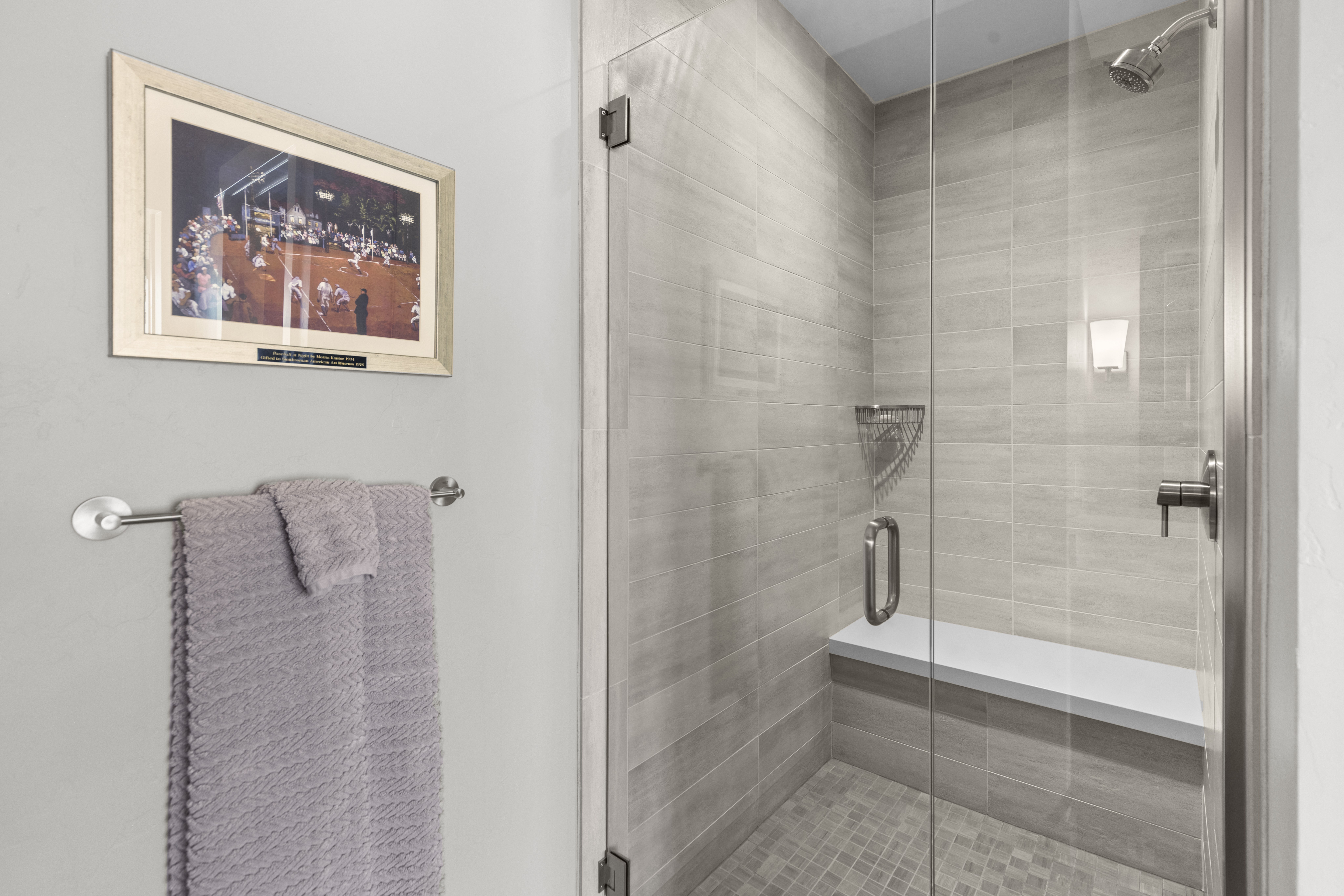 This is a well-lit bathroom featuring a modern shower with glass doors and light gray tiling. A built-in bench and a shower caddy are visible inside the shower. A framed picture hangs on the wall next to a towel rack with a neatly folded towel, adding a touch of personality to the space.