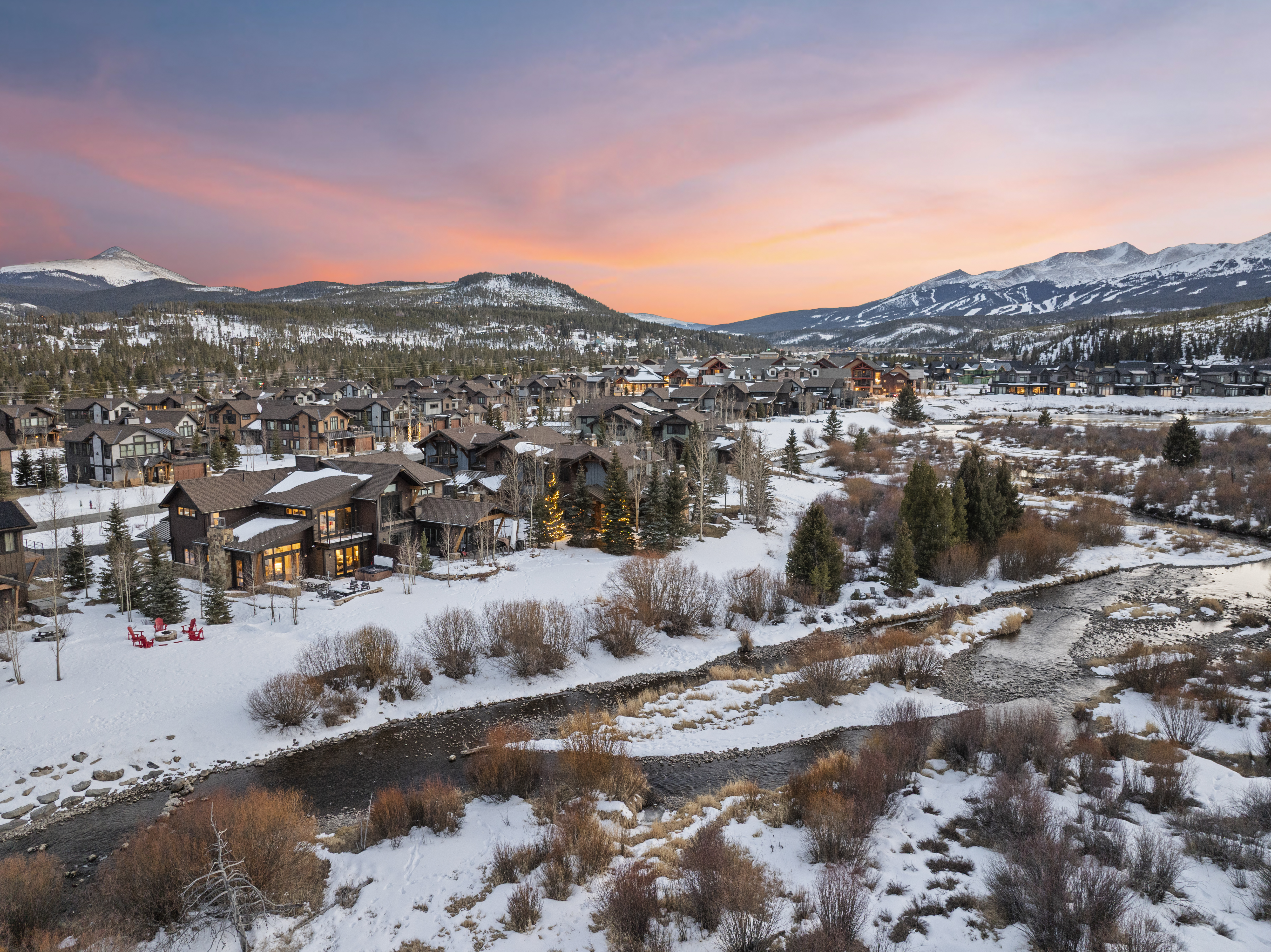 This aerial view showcases a luxurious mountain community at sunset, with snow-covered rooftops and a winding stream in the foreground. The homes feature a mix of architectural styles, blending seamlessly with the natural landscape. The pink and orange hues of the sky create a warm and inviting atmosphere, highlighting the serene beauty of this upscale neighborhood.