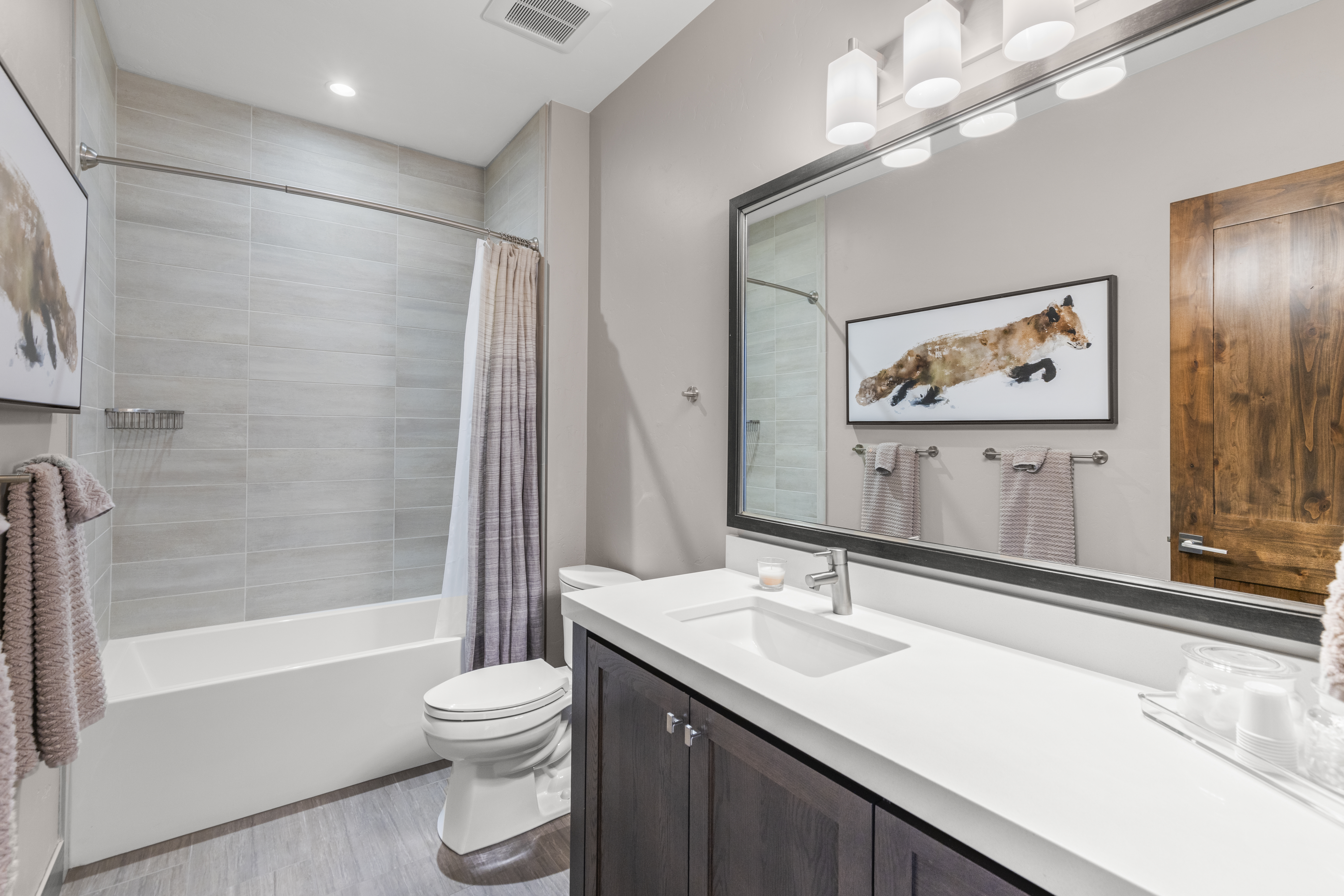 This is a well-lit bathroom featuring a white bathtub with gray tiled walls and a shower curtain. A dark wood vanity with a white countertop and a rectangular sink is present, along with a toilet. A large mirror reflects a fox artwork and a wooden door, enhancing the room's aesthetic appeal.
