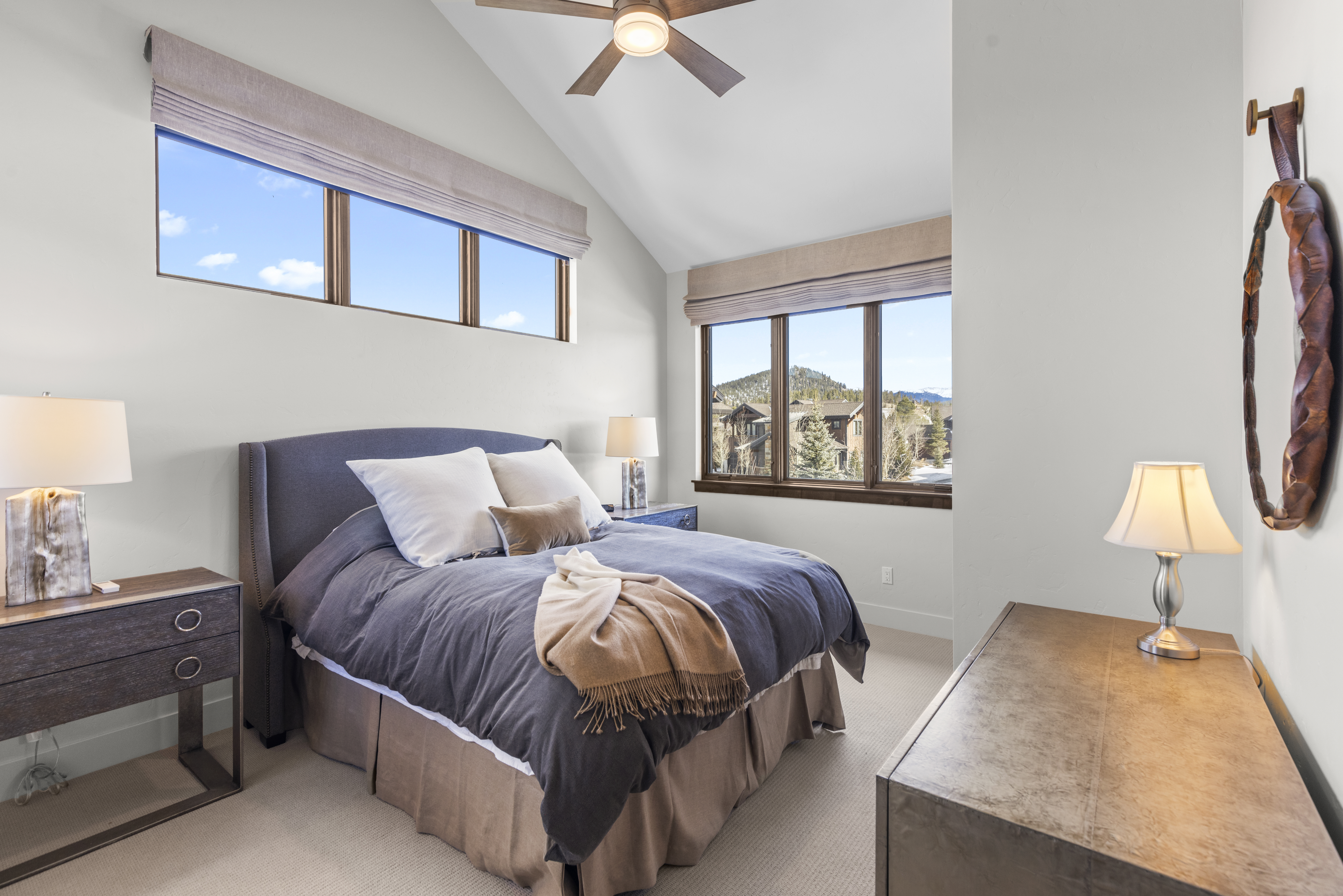 This is a primary bedroom featuring a large bed with a gray headboard and a blue comforter. The room has two windows with views of the outdoors, and the walls are painted a neutral color. There are two nightstands with lamps on either side of the bed, and a dresser is visible on the right side of the frame. The perspective is from the foot of the bed, looking towards the windows.