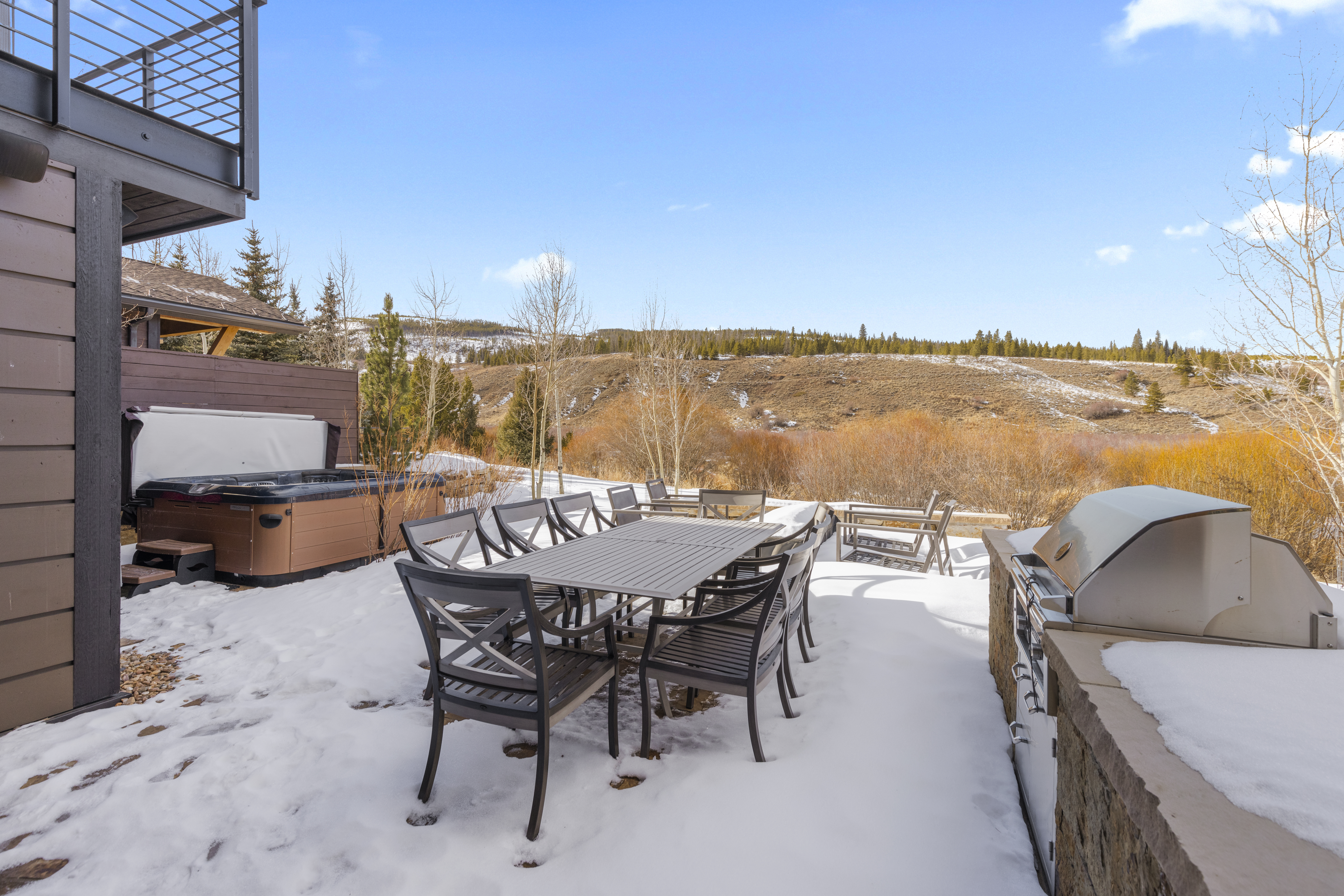 This is an exterior shot of a patio or deck area, covered in snow. The space features a large outdoor dining table with chairs, a hot tub, and a built-in grill area. The background showcases a scenic view of rolling hills and sparse vegetation, suggesting a mountain or rural setting.