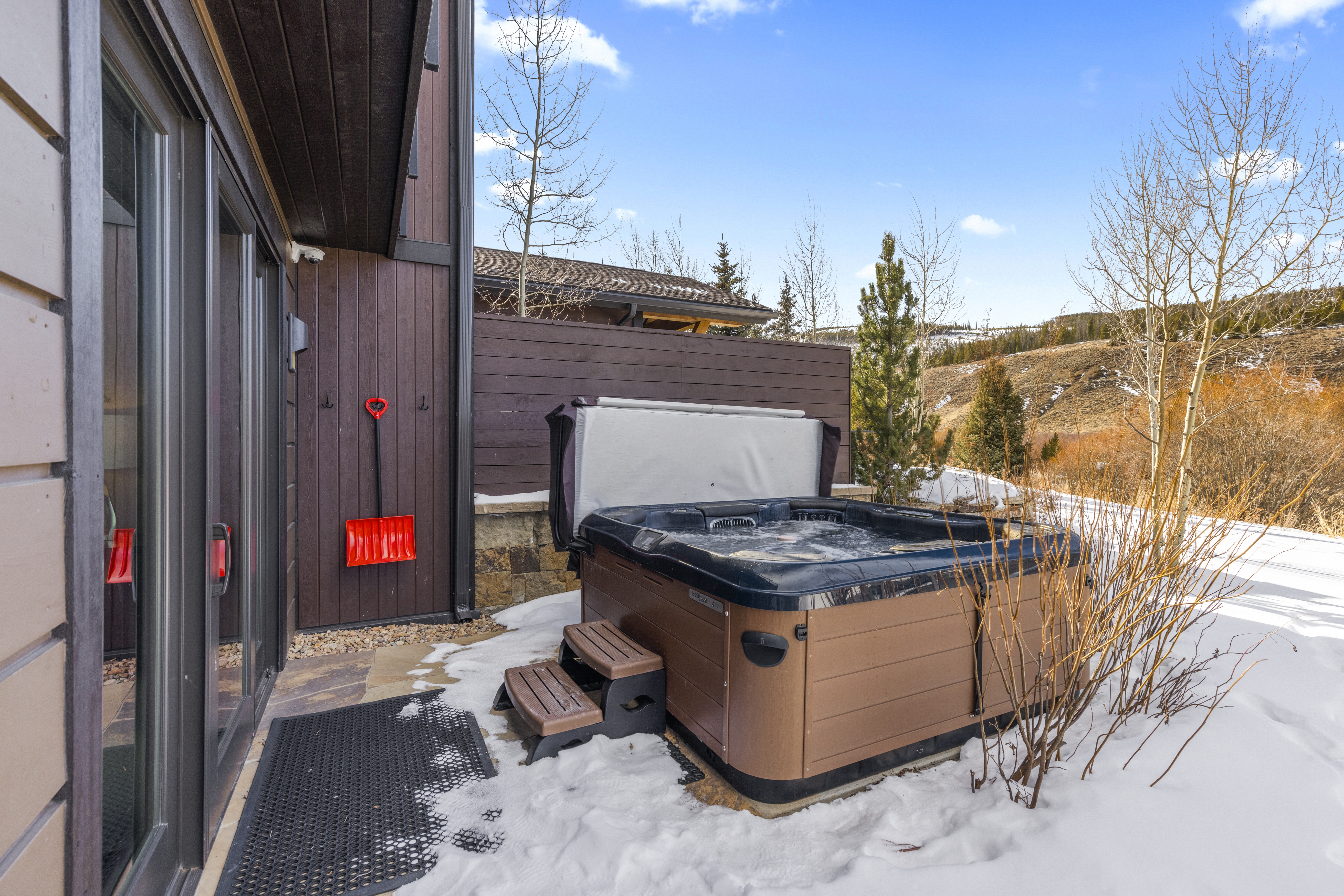 This image showcases a private outdoor patio area featuring a hot tub, perfect for relaxation and entertainment. The patio is partially covered in snow, suggesting a winter setting, and is adjacent to the building with easy access through a sliding glass door. A red snow shovel is hung on the wall, adding a practical touch.