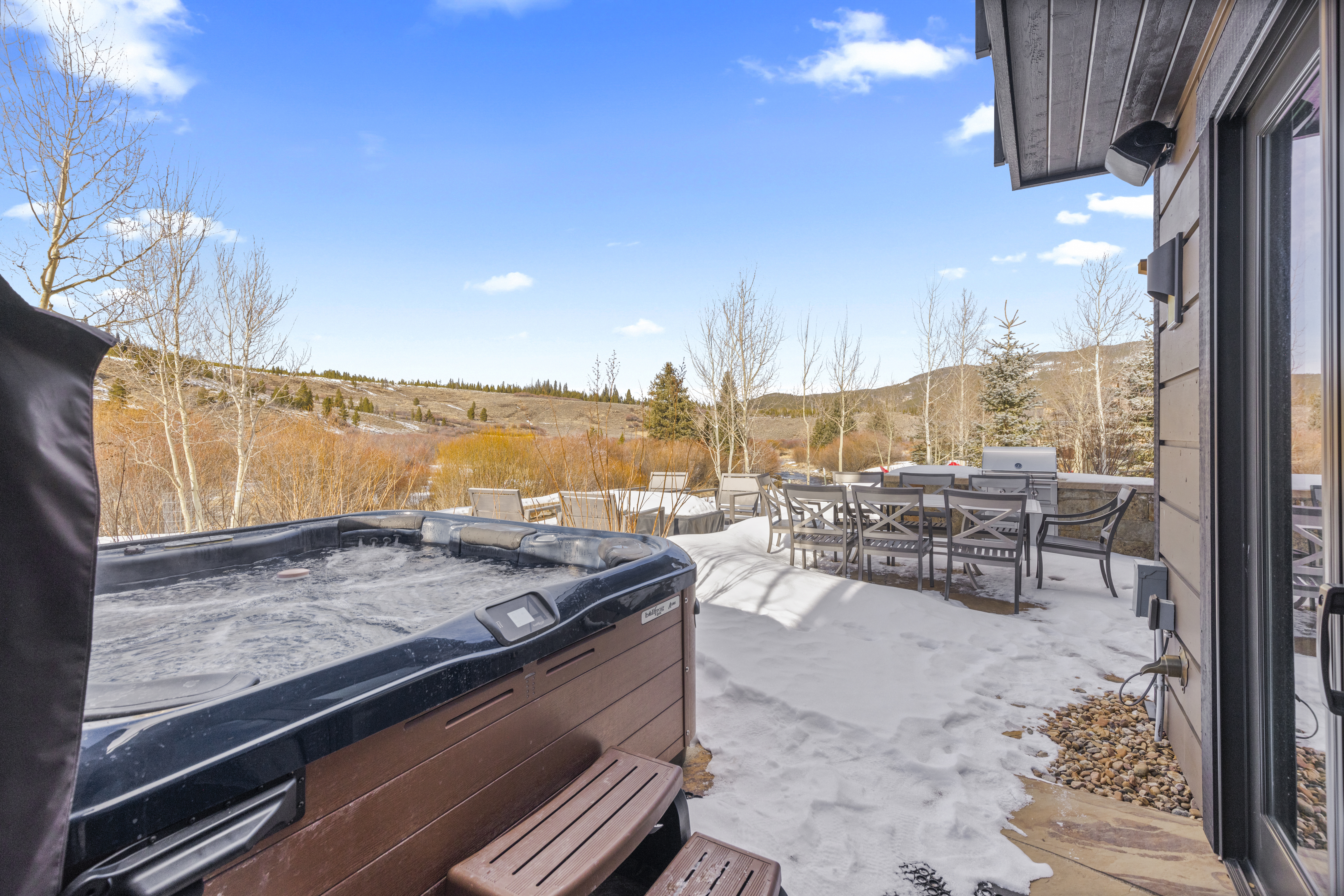 This image showcases a patio or deck area featuring a hot tub and outdoor dining set. The patio is partially covered in snow, suggesting a mountain or colder climate setting. The dining set includes a table and chairs, and a grill is visible in the background, indicating an outdoor cooking area. The hot tub is a prominent feature, suggesting relaxation and leisure.