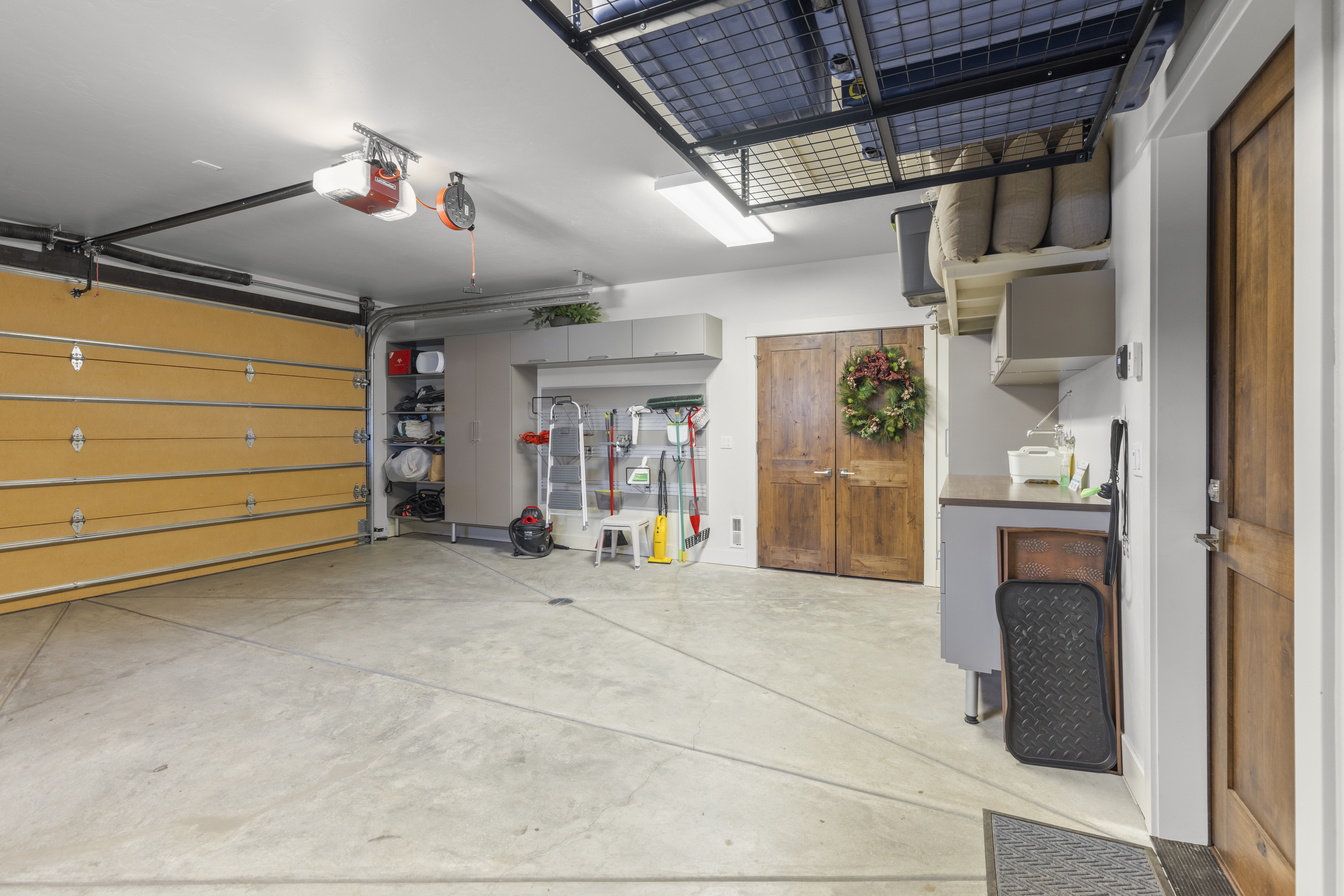 This is an interior shot of a well-organized garage. The garage features a light-colored concrete floor, a wooden garage door, and built-in storage cabinets along the back wall. There is also a storage rack suspended from the ceiling, and a door leading to the interior of the house.