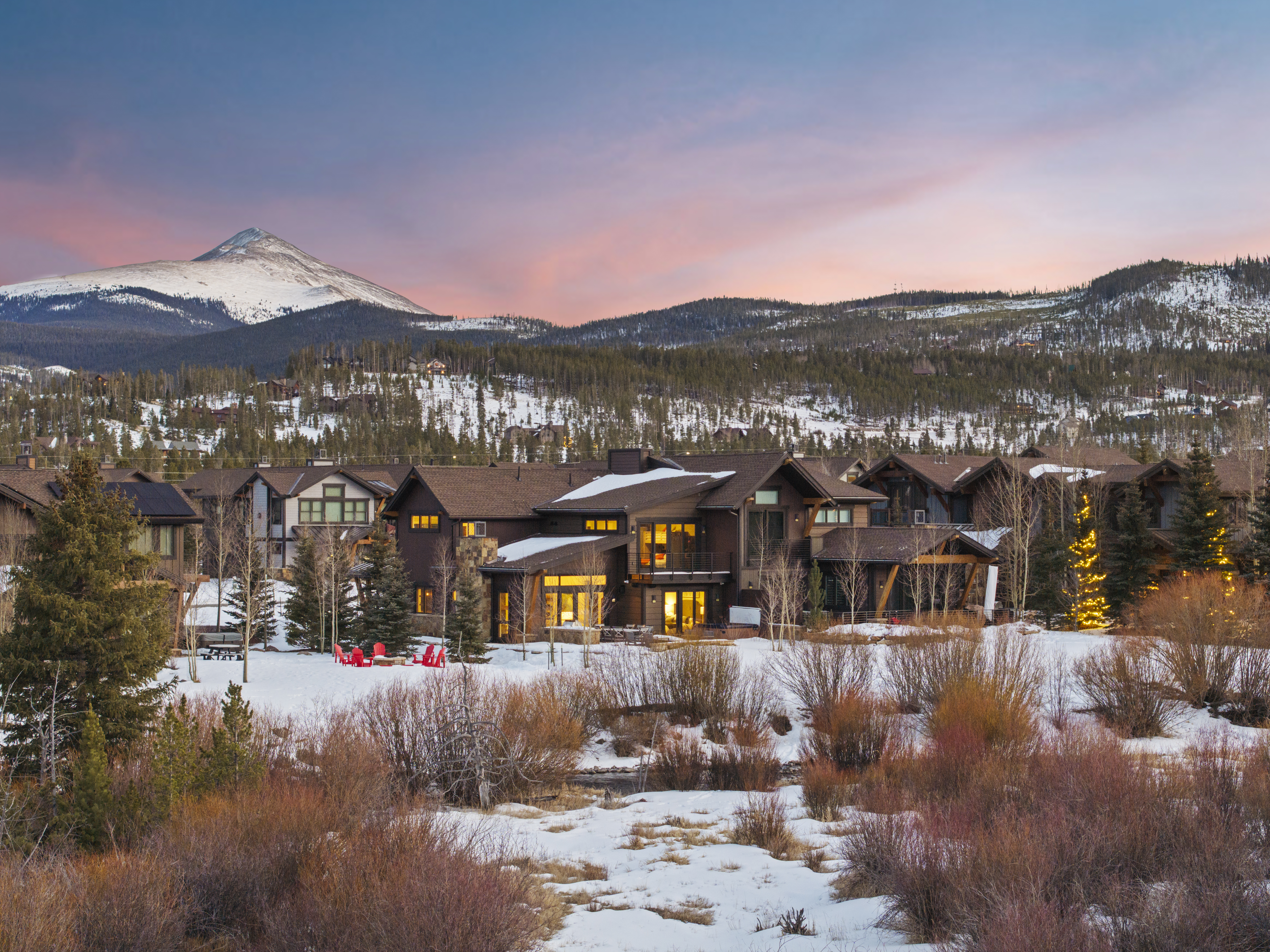 This image showcases the front exterior of a luxurious mountain home at dusk. The architecture features a blend of natural wood and stone elements, with warm interior lighting visible through the windows. The property is surrounded by a snow-covered landscape with mature trees and a mountain backdrop, creating a serene and inviting atmosphere.