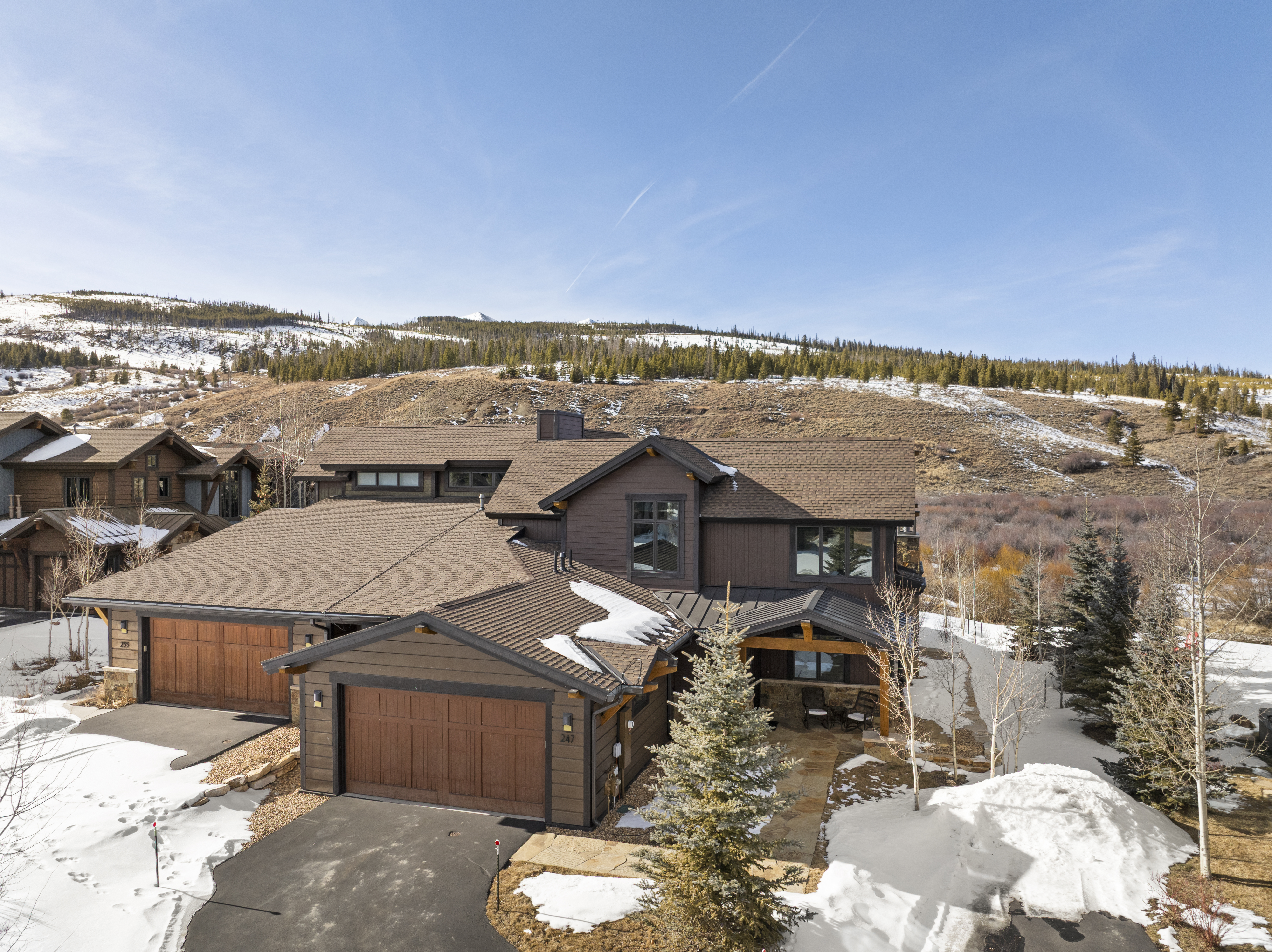 This aerial view showcases a luxurious mountain home with a brown exterior and a matching roof. The property features a multi-car garage, a covered entryway, and is surrounded by a landscape that blends natural terrain with snow-covered areas. The home exudes a sense of rustic elegance and privacy.
