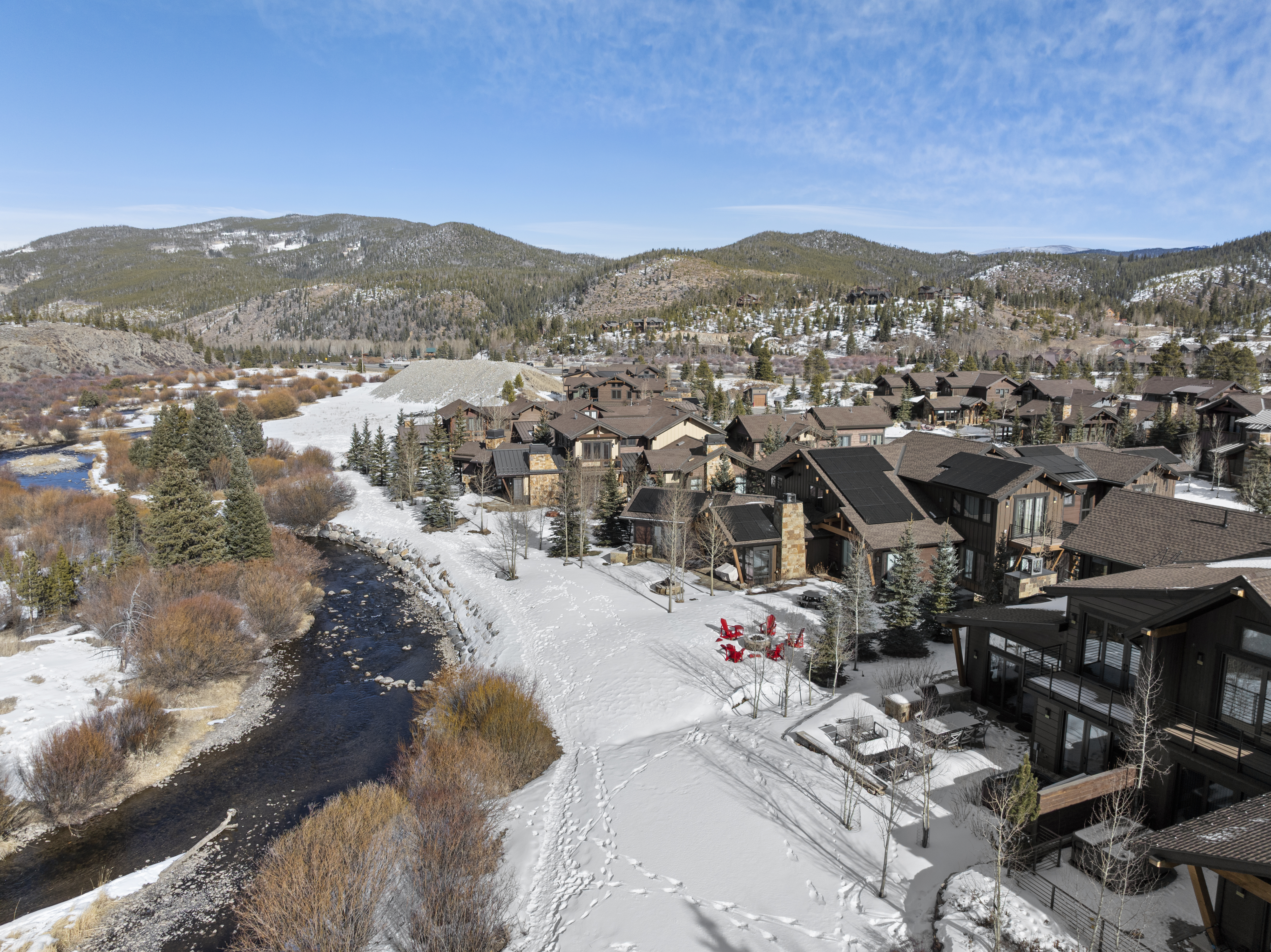 This aerial view showcases a luxurious residential community nestled in a snowy mountain landscape. The homes feature a mix of stone and wood exteriors with dark roofs, blending harmoniously with the natural surroundings. A winding river adds a serene touch to the scene, enhancing the property's appeal and creating a sense of tranquility and exclusivity.