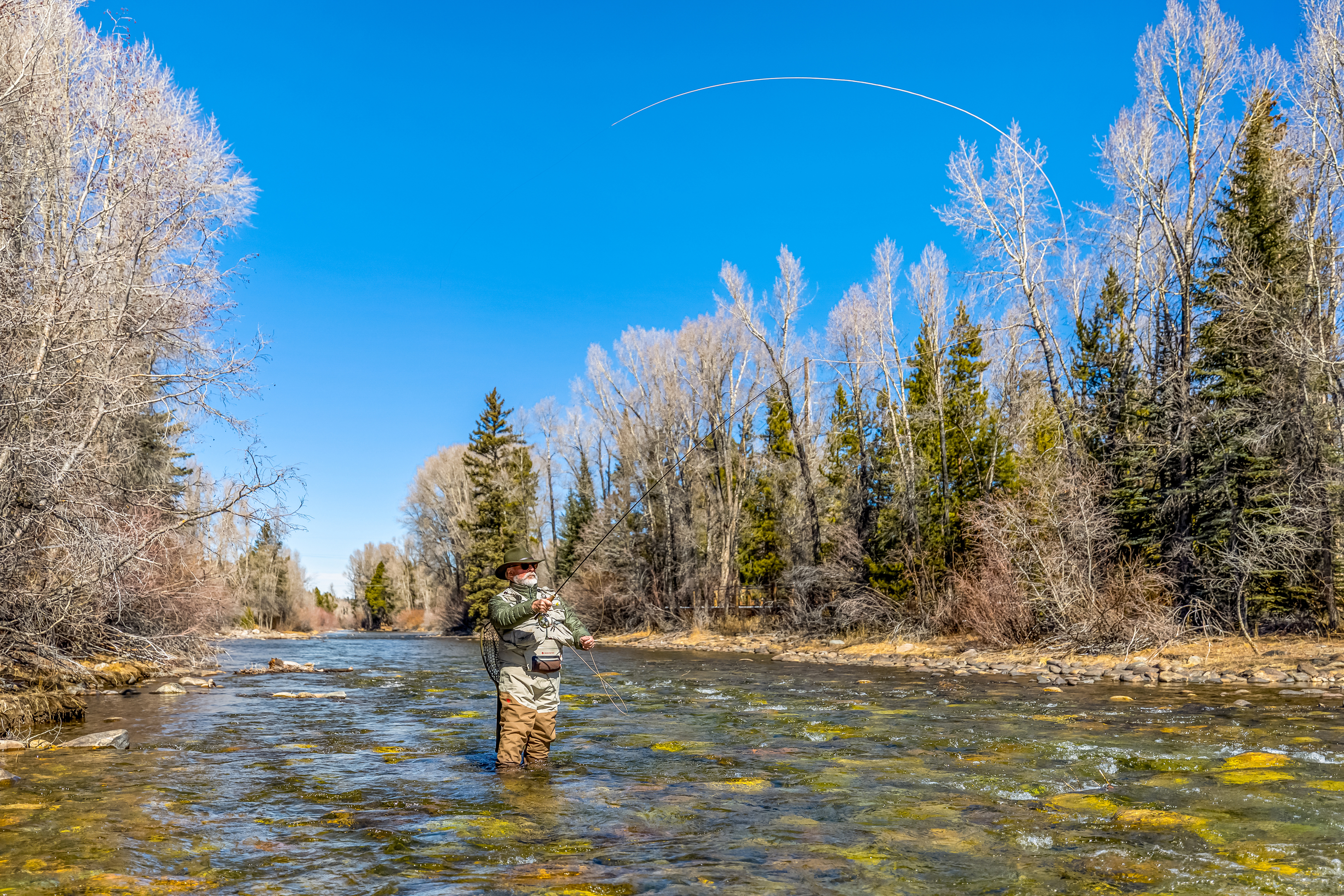 This image captures a serene, wide-angle shot of a fly fisherman standing in the middle of a clear, shallow river, casting his line into the air against a backdrop of bare, late-autumn trees and a vibrant blue sky. The scene emphasizes the natural beauty and recreational potential of the surrounding landscape, highlighting the tranquil environment and clear water conditions. The perspective is eye-level, drawing the viewer into the peaceful, outdoor setting.