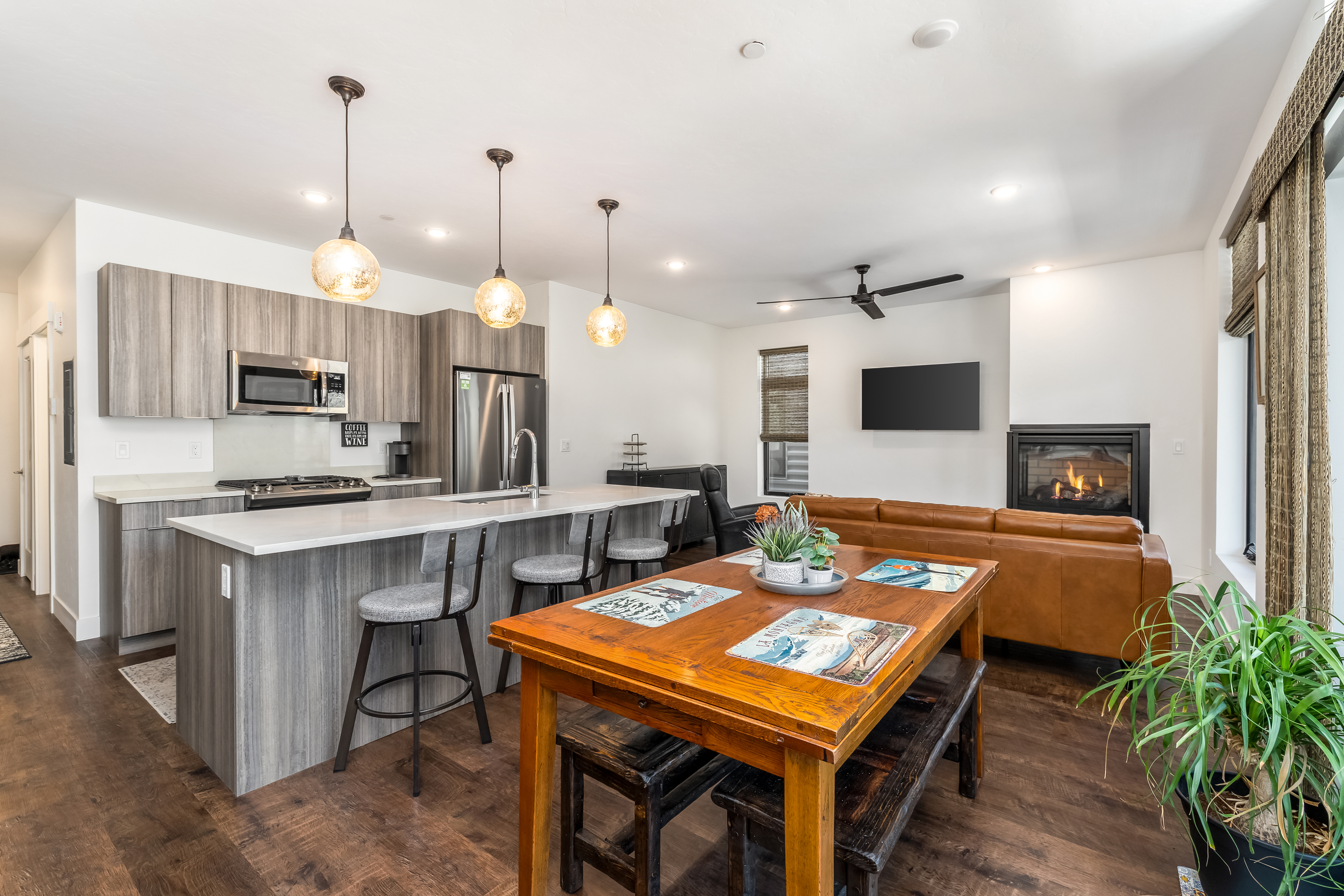 This open-concept living space features a modern kitchen with sleek wood-grain cabinetry, stainless steel appliances, and a spacious island with bar seating. The area flows seamlessly into a dining space with a rustic wooden table and a cozy living room anchored by a fireplace and a leather sofa. The warm wood flooring and contemporary lighting create an inviting and functional atmosphere perfect for entertaining.