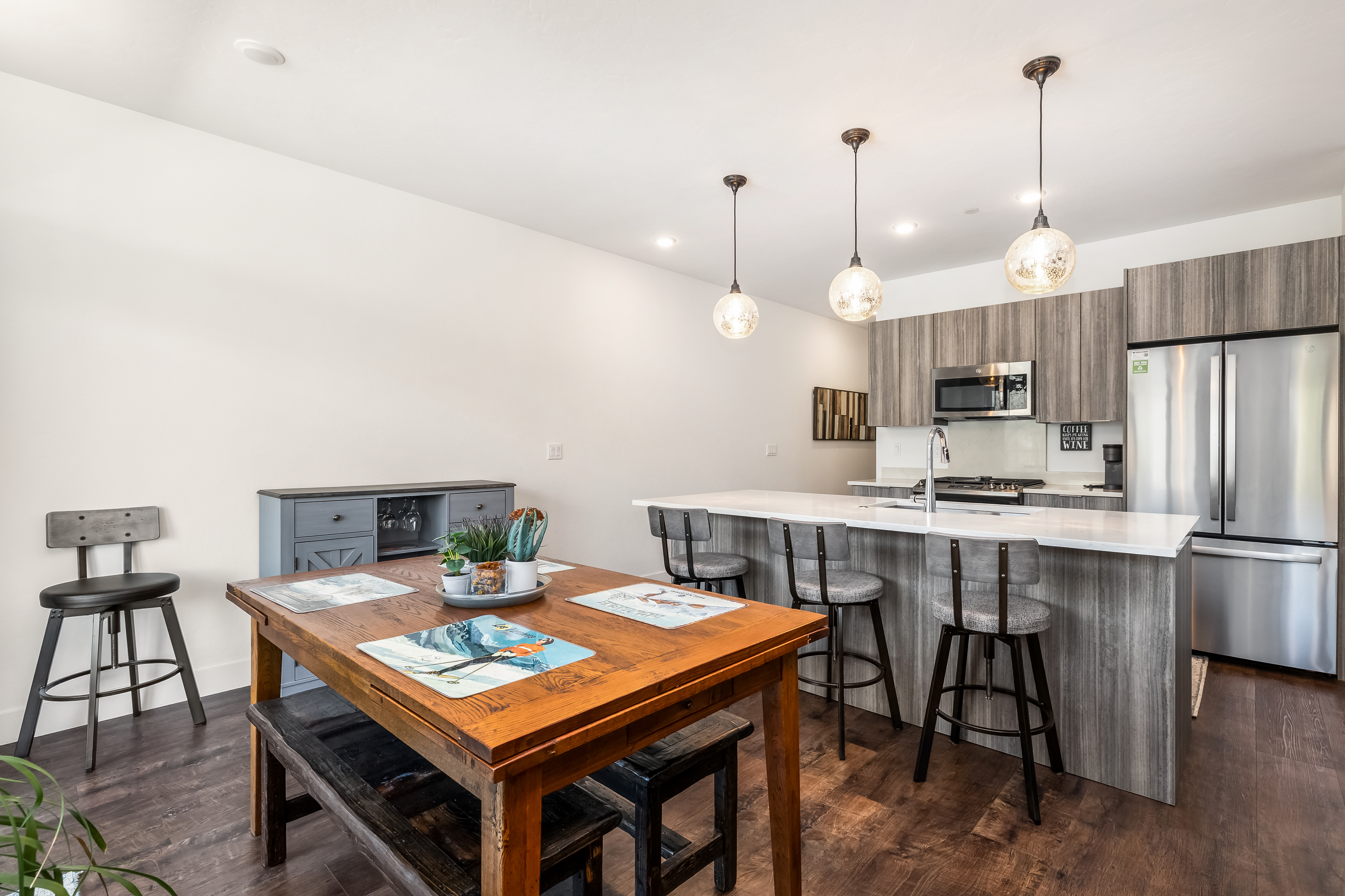 This open-concept kitchen and dining area features a rustic wooden dining table paired with a bench and stools, complemented by a modern kitchen island with bar seating. The space is defined by sleek, grey-toned cabinetry, stainless steel appliances, and elegant pendant lighting that creates a warm, inviting atmosphere. The combination of industrial-style stools and natural wood textures provides a contemporary yet cozy aesthetic perfect for entertaining.