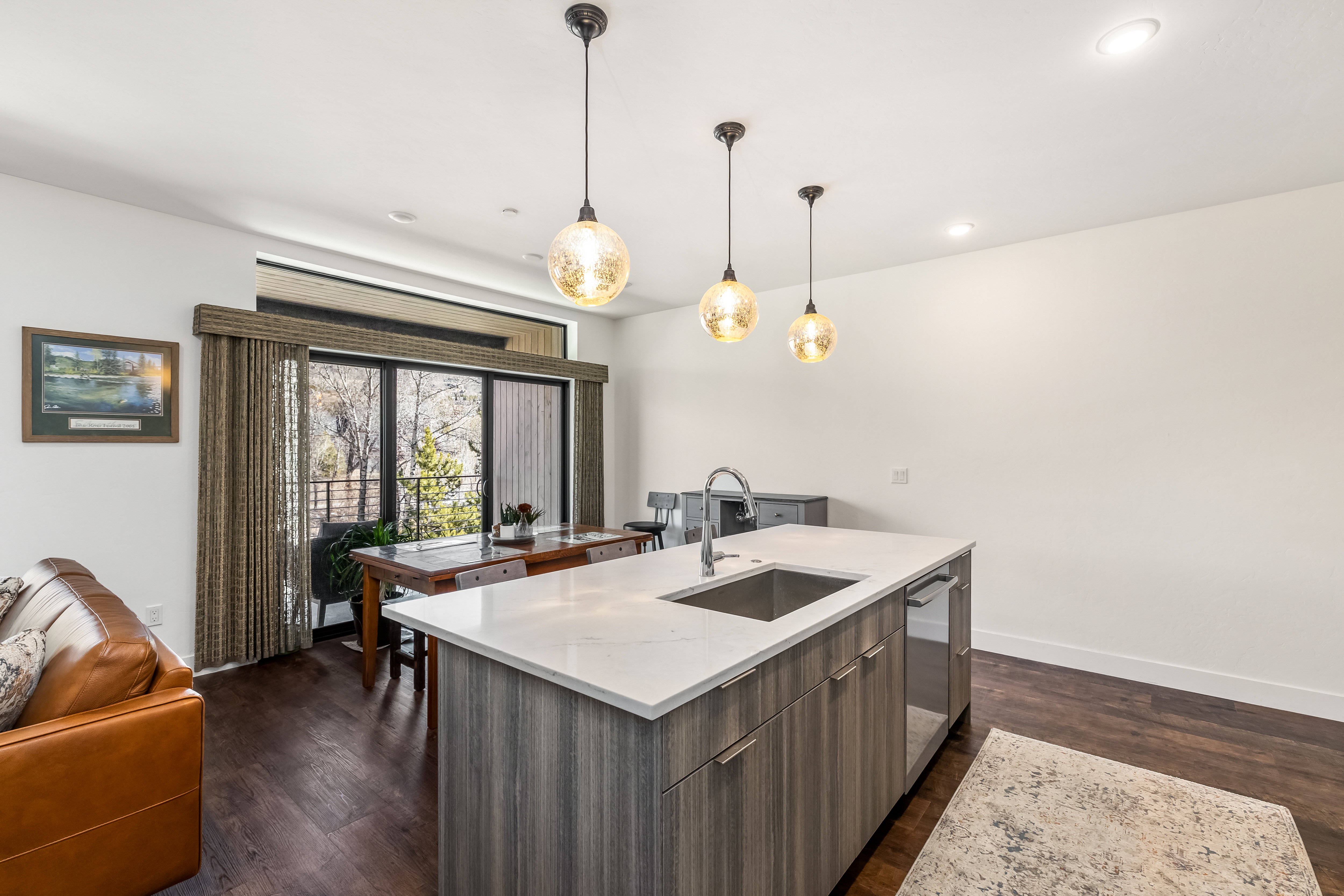 This modern kitchen features a large island with a quartz countertop, a stainless steel sink, and a built-in dishwasher, all set against a backdrop of wood-grain cabinetry. Three elegant, textured glass pendant lights hang above the island, while a dining area with a wooden table is positioned near a large sliding glass door that leads to a balcony. The space is bright and open, showcasing a contemporary design with dark hardwood flooring and a neutral color palette.