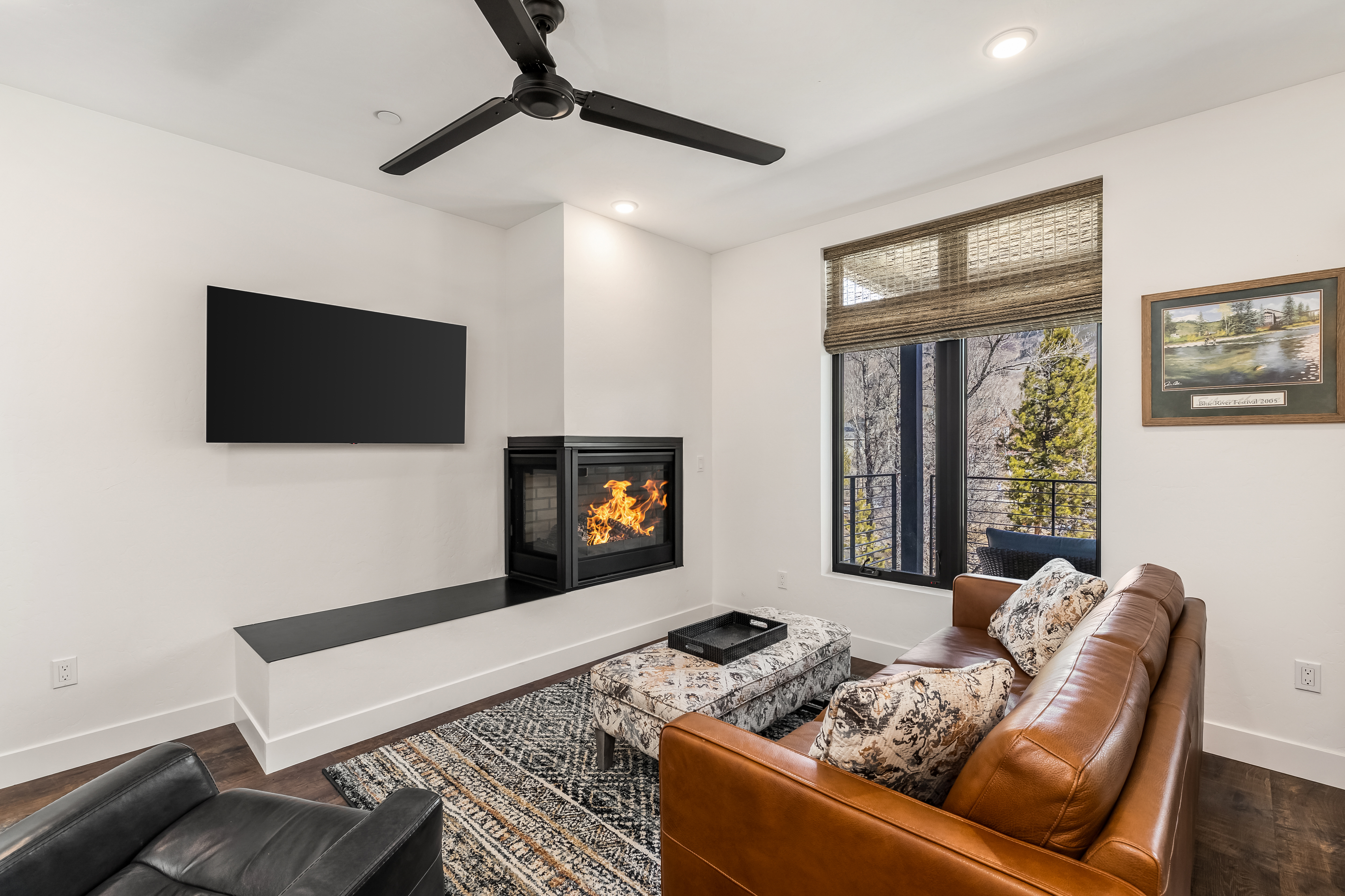 This cozy and modern living room features a warm leather sofa, a patterned ottoman, and a sleek corner fireplace that serves as a focal point. A large flat-screen television is mounted on the wall above a minimalist hearth, while a window with woven shades provides a view of the outdoors. The space is anchored by a textured area rug and illuminated by a contemporary ceiling fan, creating an inviting and comfortable atmosphere.