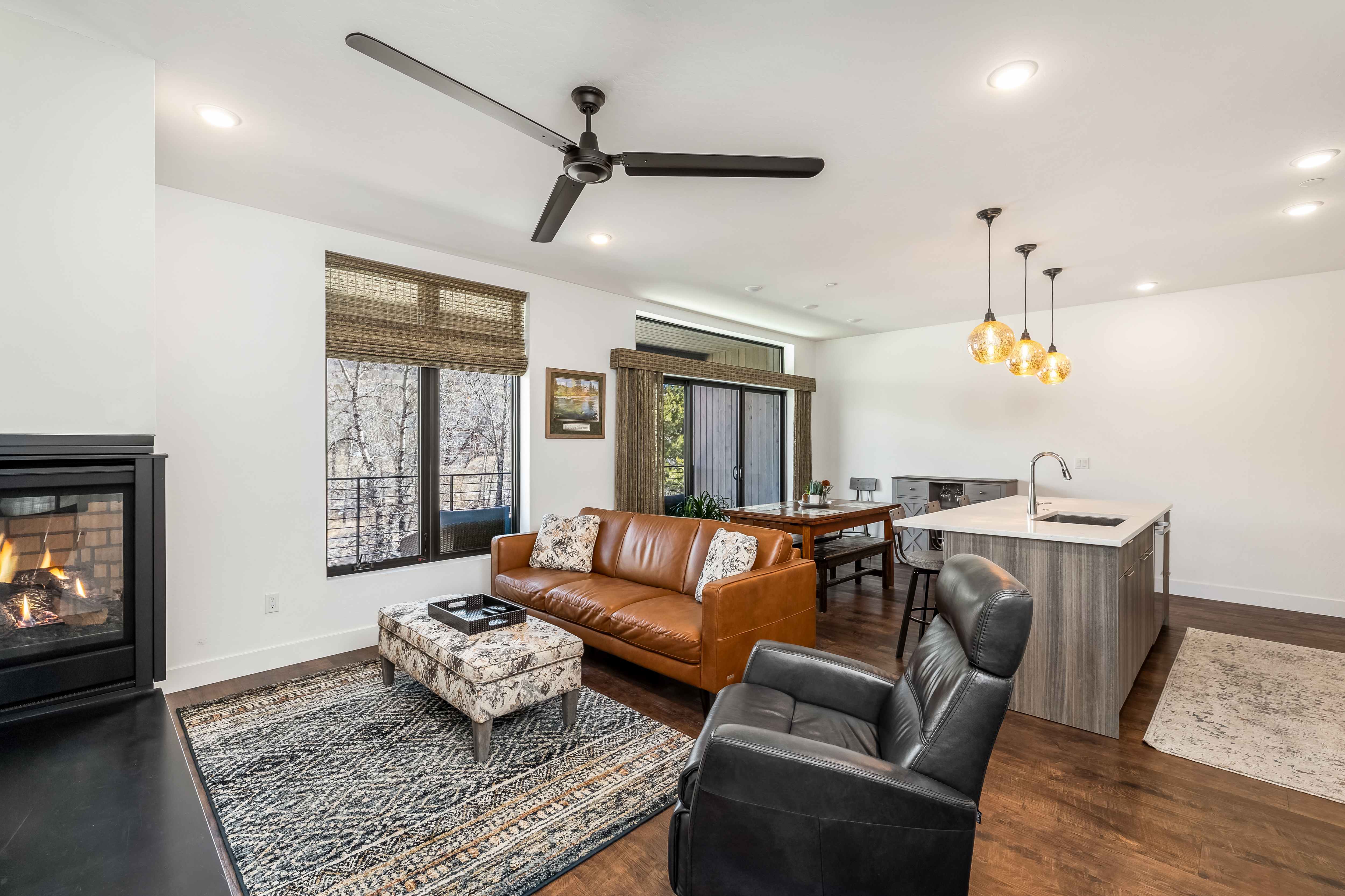 This open-concept living space features a warm, inviting aesthetic with a rich brown leather sofa, a patterned ottoman, and a sleek black leather recliner. The room is anchored by a modern fireplace on the left and flows seamlessly into a dining area and a contemporary kitchen with a large island. Large windows with woven shades provide natural light and a view of the outdoors, creating a comfortable and cohesive living environment.