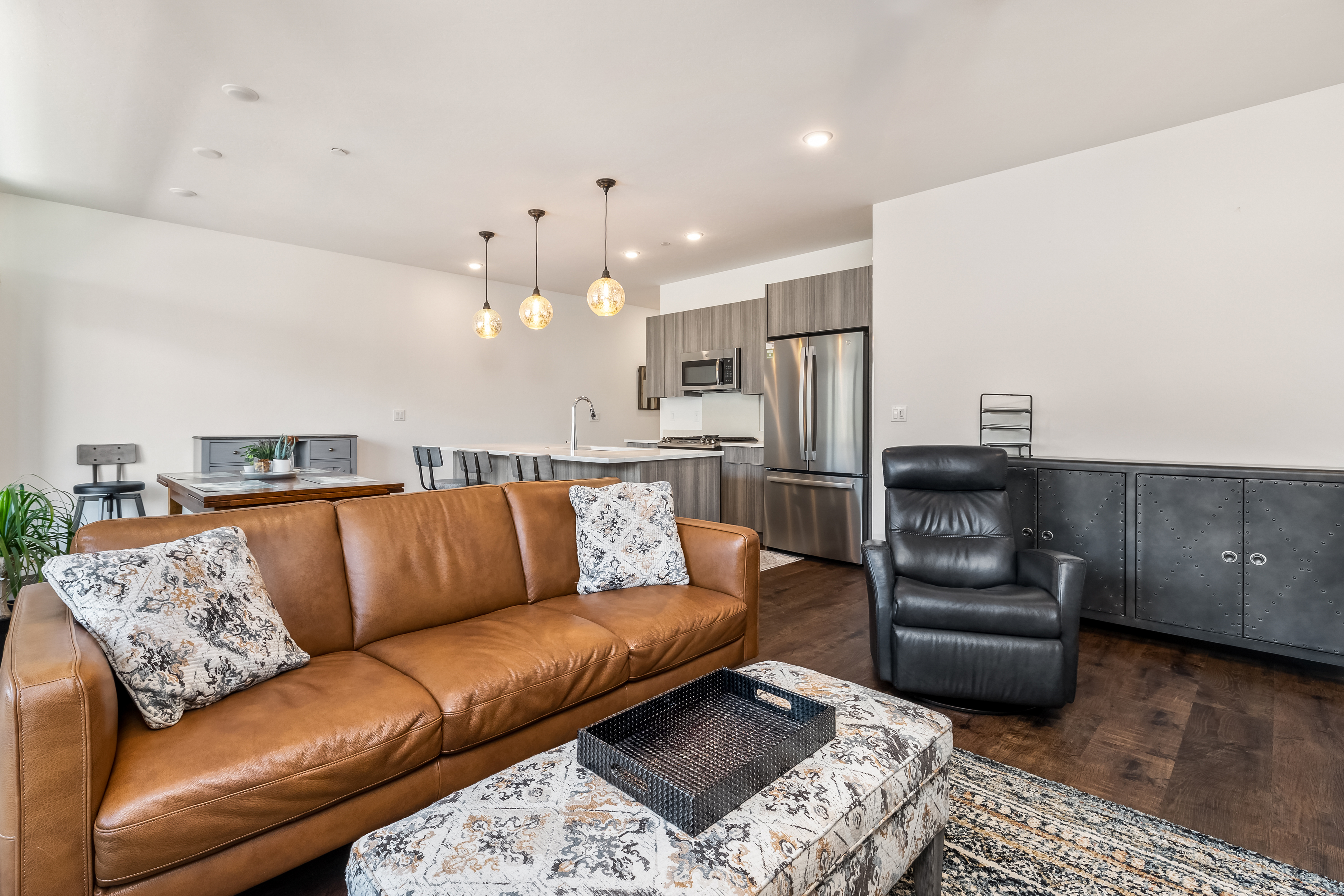 This open-concept living space features a warm, inviting brown leather sofa paired with a patterned ottoman and a sleek black leather recliner. The room flows seamlessly into a modern kitchen with stainless steel appliances, dark wood cabinetry, and a breakfast bar illuminated by three pendant lights. The dark hardwood flooring and industrial-style metal sideboard create a sophisticated, contemporary aesthetic perfect for comfortable living and entertaining.
