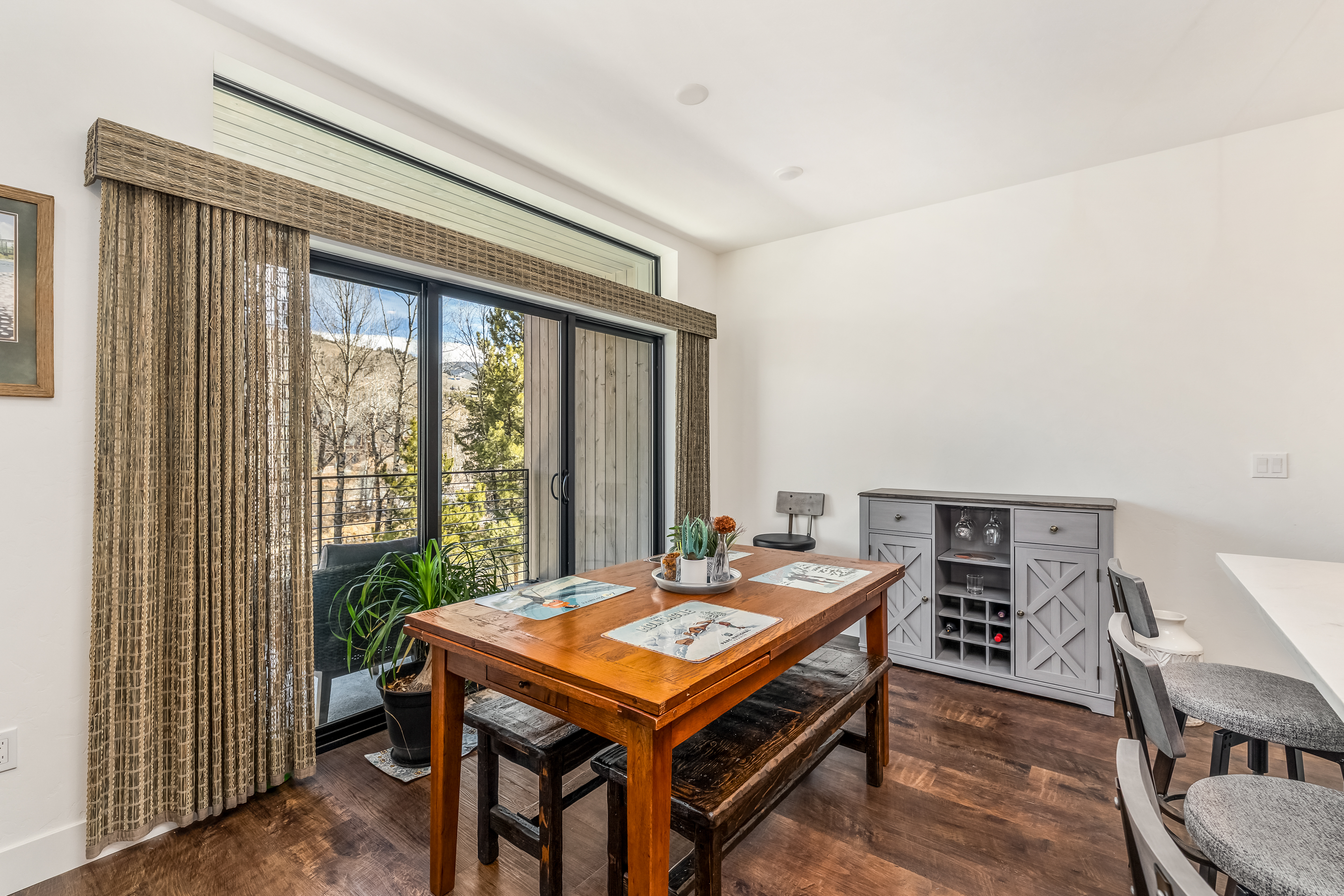 This inviting dining area features a rustic wooden table paired with matching benches, set against a backdrop of large sliding glass doors that offer a scenic view of the outdoors. A grey sideboard with built-in wine storage adds a touch of modern functionality, while the warm wood tones and natural light create a cozy, mountain-inspired atmosphere. The perspective is eye-level, capturing the seamless transition between the indoor dining space and the adjacent balcony.