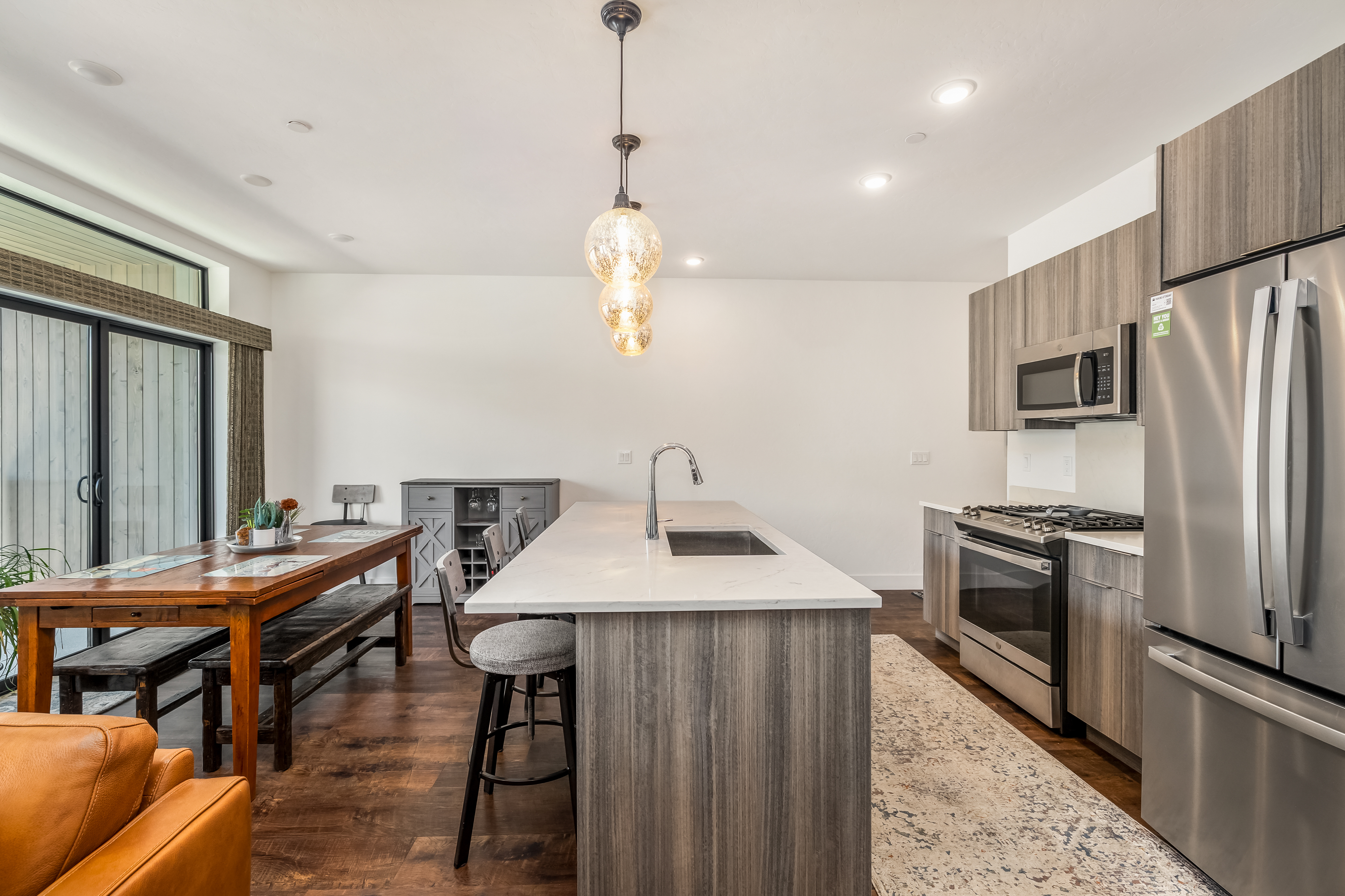 This modern open-concept kitchen features a large island with a quartz countertop, stainless steel appliances, and sleek wood-grain cabinetry. The space seamlessly transitions into a dining area with a rustic wooden table and bench seating, all illuminated by elegant glass pendant lights. The warm wood flooring and neutral color palette create a sophisticated and inviting atmosphere perfect for entertaining.