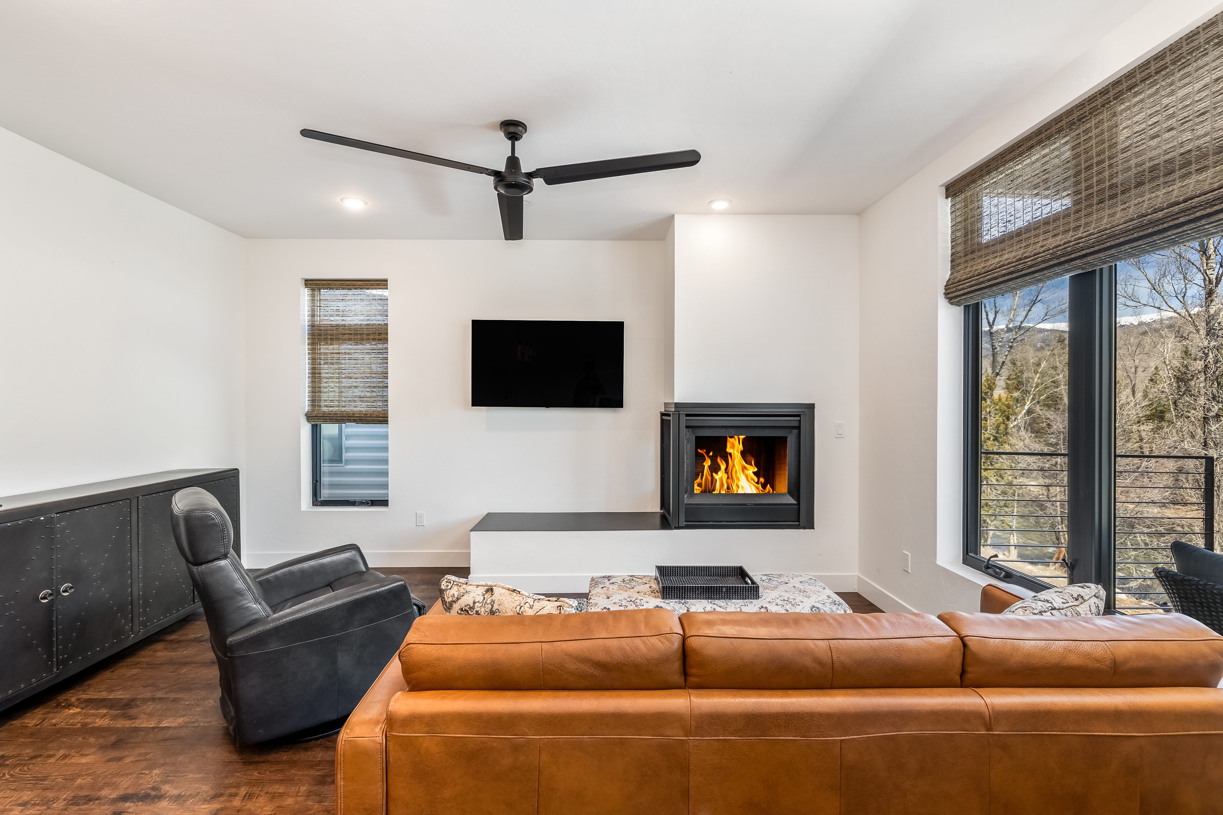 This modern living room features a warm, inviting aesthetic with a large cognac-colored leather sofa and a sleek black leather recliner. The focal point is a contemporary fireplace integrated into a minimalist white wall, positioned next to a wall-mounted television and a large window offering a scenic outdoor view. The space is grounded by rich dark hardwood flooring and a ceiling fan, creating a sophisticated yet comfortable atmosphere.