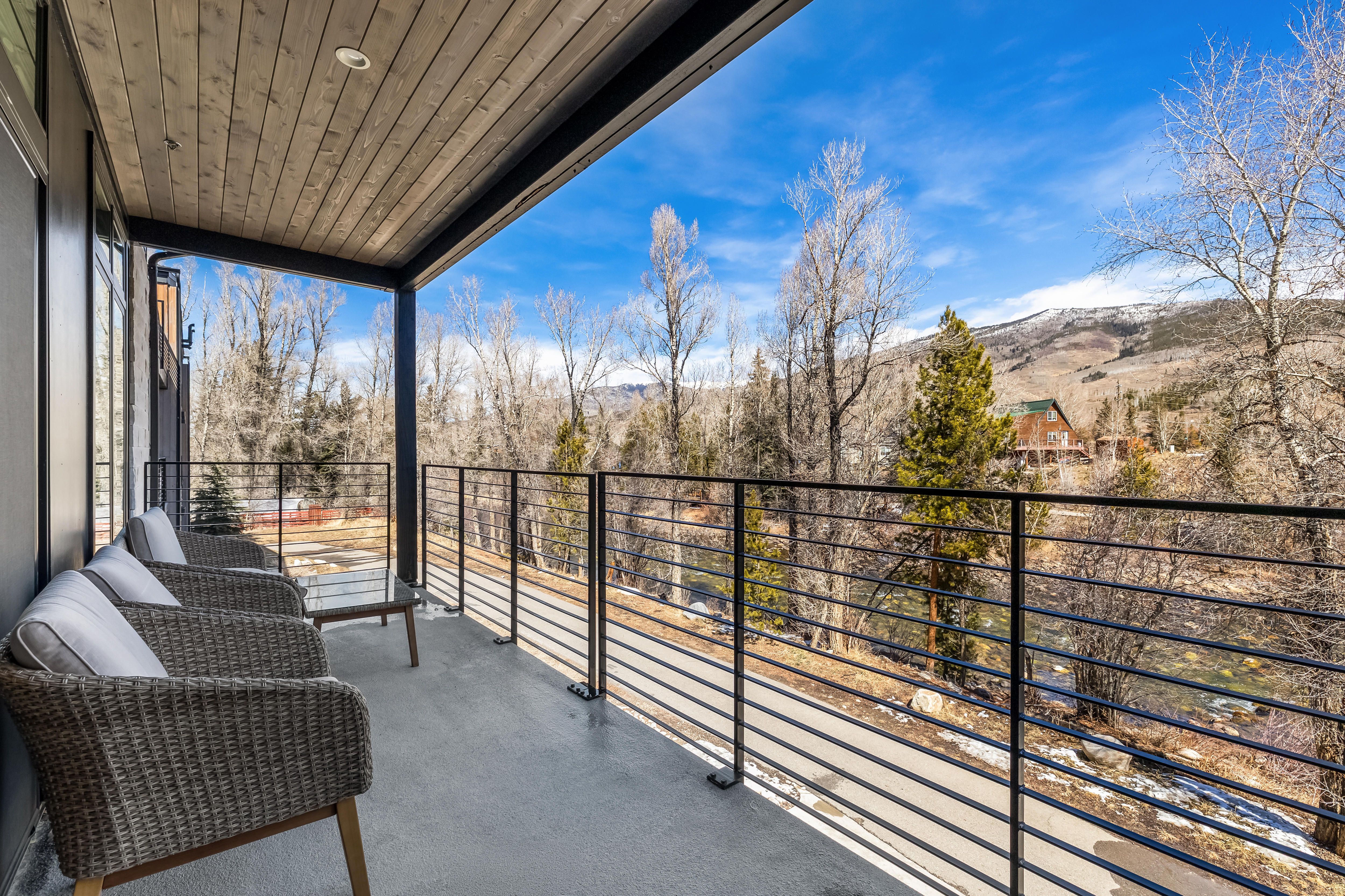 This modern balcony features sleek, minimalist metal railings and a covered ceiling with recessed lighting, offering a sophisticated outdoor living space. The area is furnished with comfortable wicker chairs and a small side table, perfectly positioned to enjoy the scenic view of the river, surrounding trees, and distant mountains. The perspective is from the edge of the balcony, emphasizing the seamless transition between the contemporary interior design and the natural landscape.